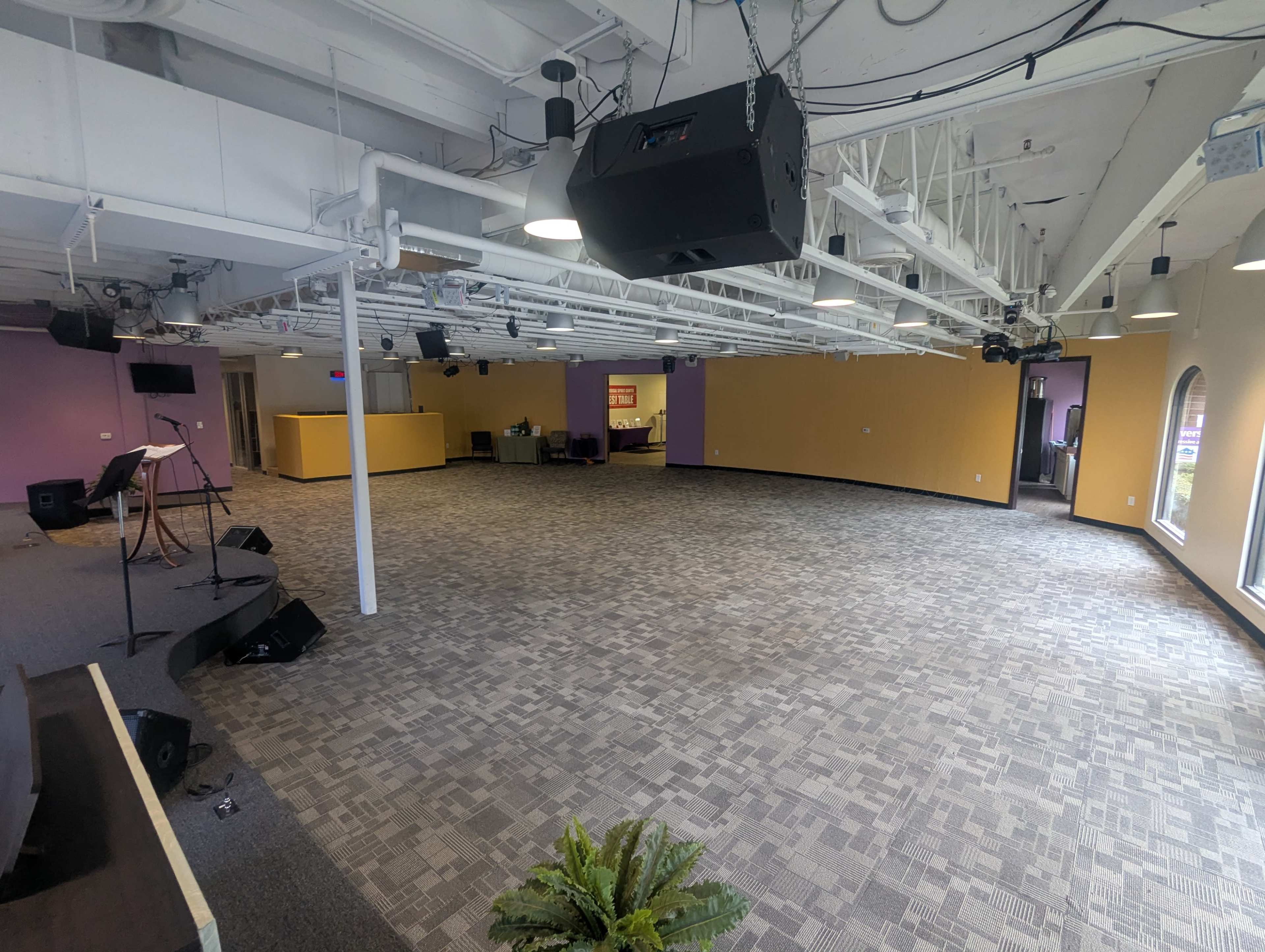 The image shows a spacious indoor area with a carpeted floor, purple and yellow walls, and a staging area for performances or presentations.