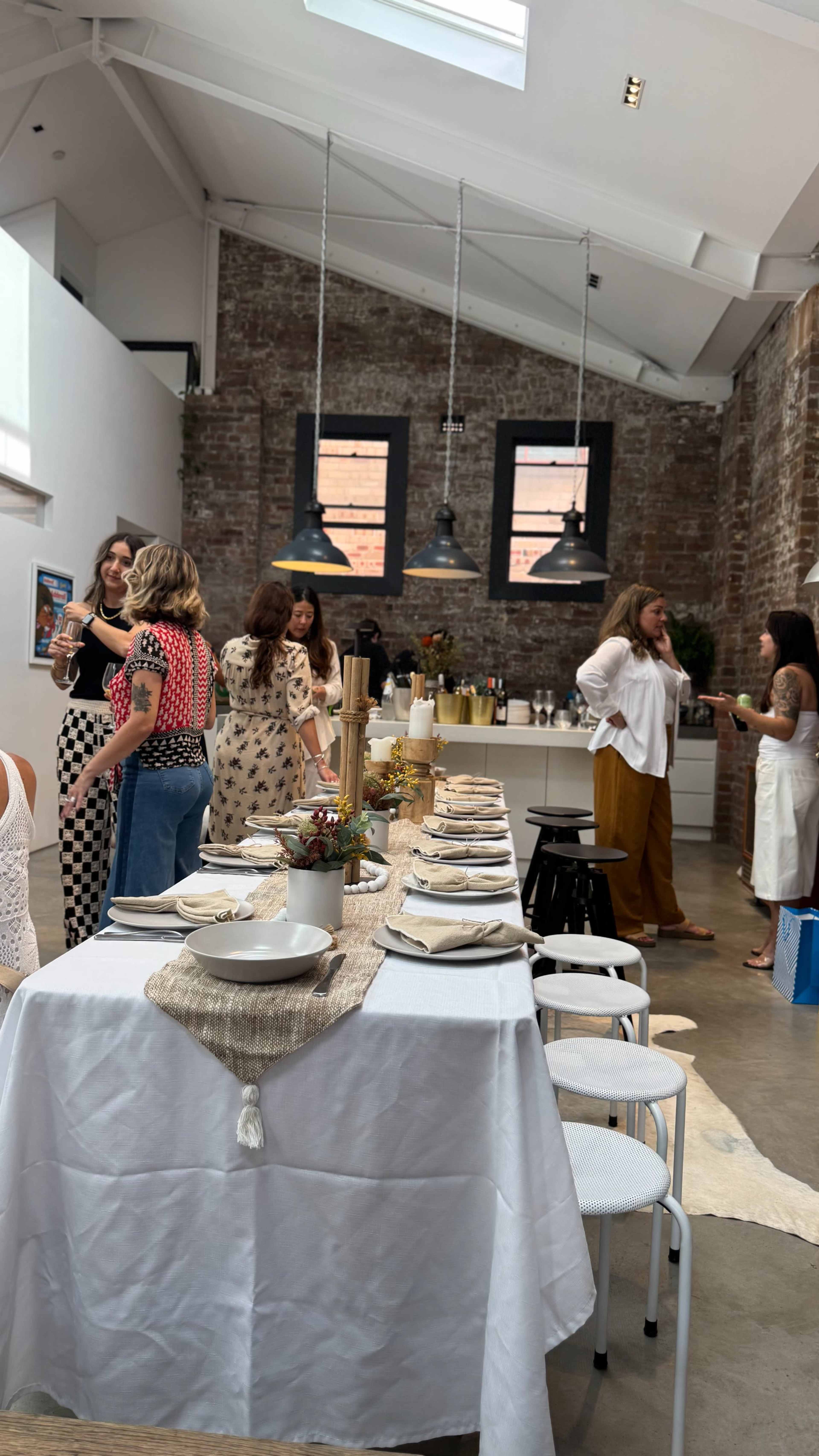 A group of women interacts in a stylish, open kitchen space with an elegantly set dining table in the foreground.
