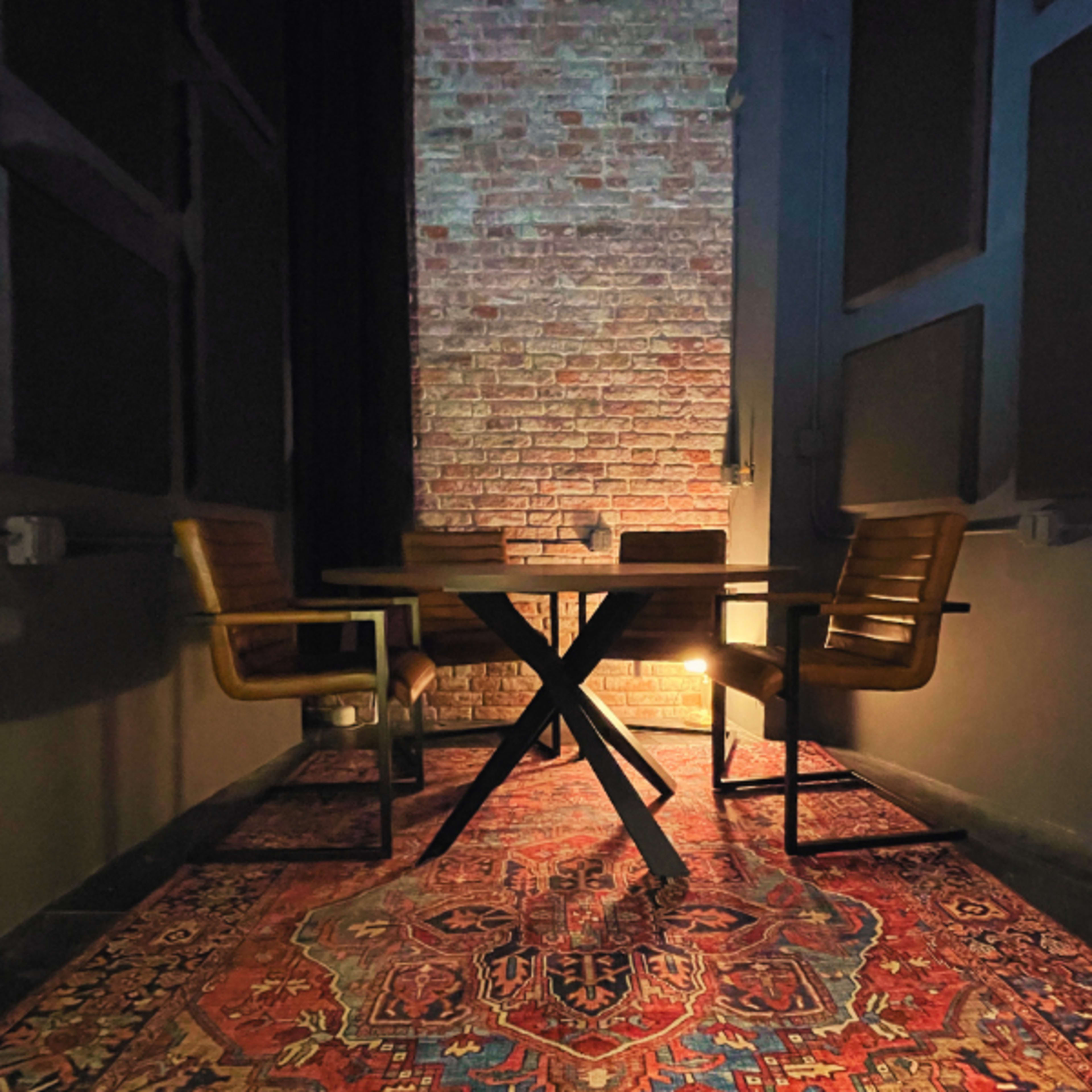 A small meeting room features a round table surrounded by four wooden chairs, set against a brick wall with soft lighting illuminating a patterned rug.