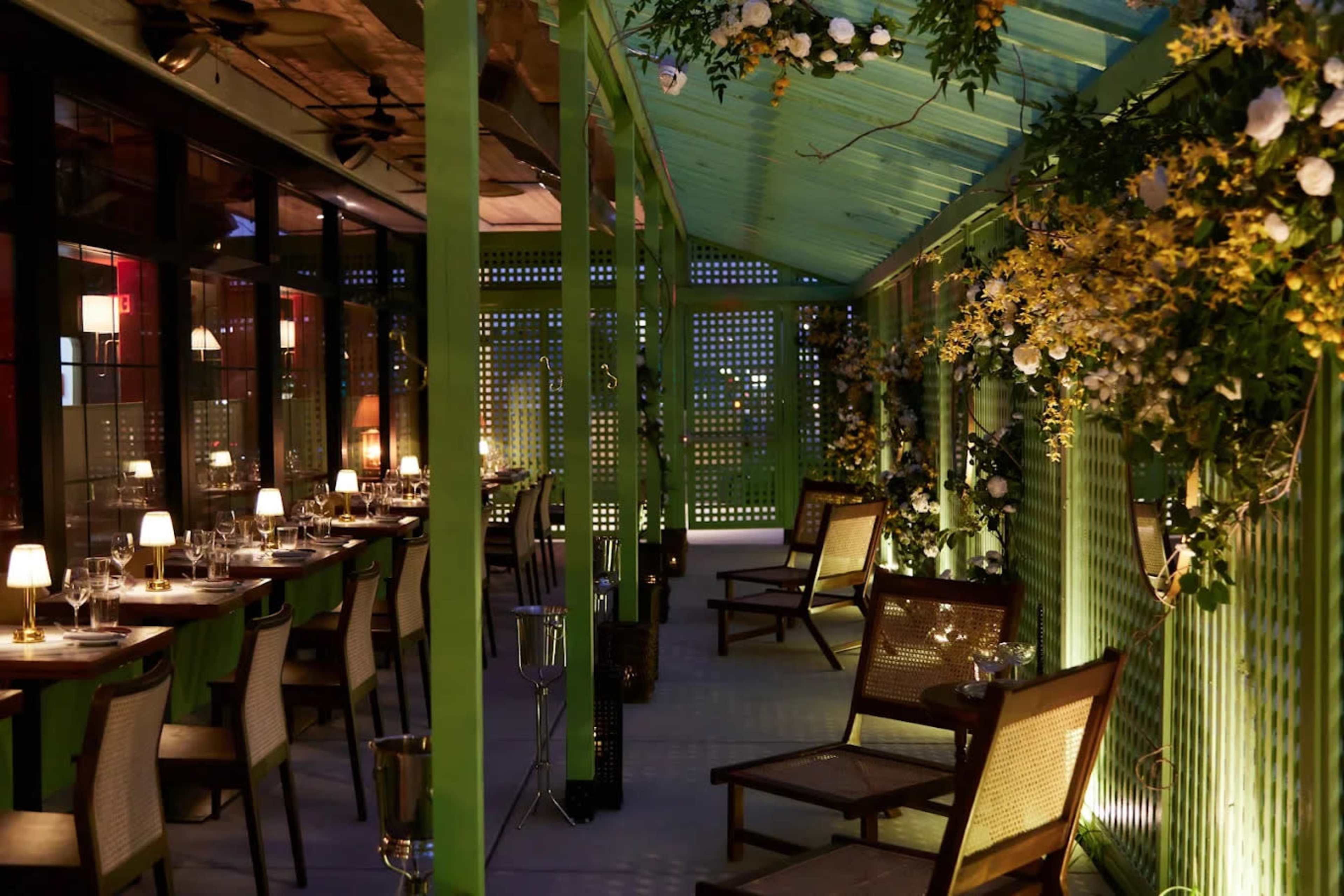 The image shows a beautifully lit indoor patio with wooden tables, chairs, and decorative flowers along the walls.