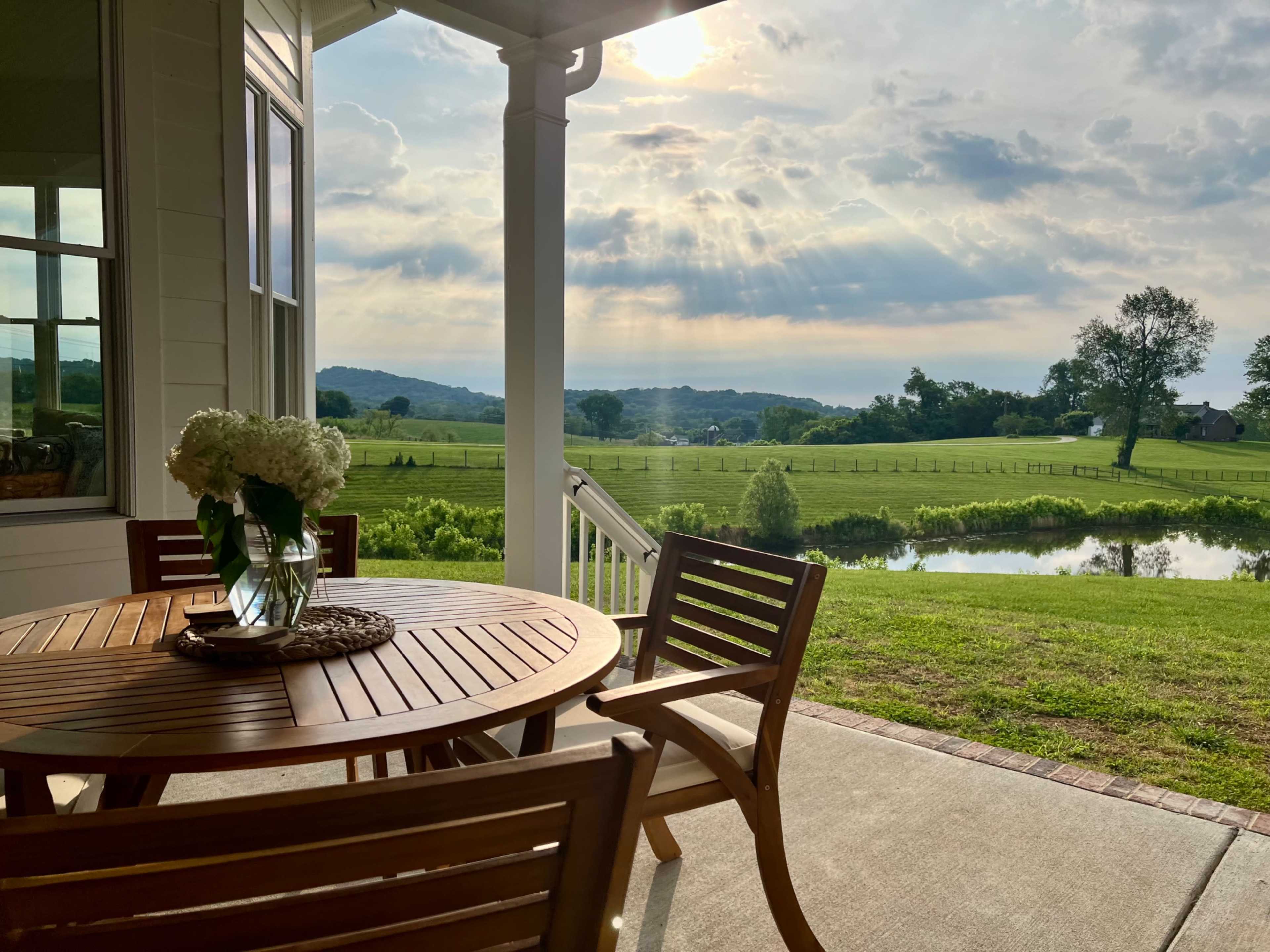 A wooden table and chairs are set on a porch overlooking a lush green landscape with rolling hills and a pond.