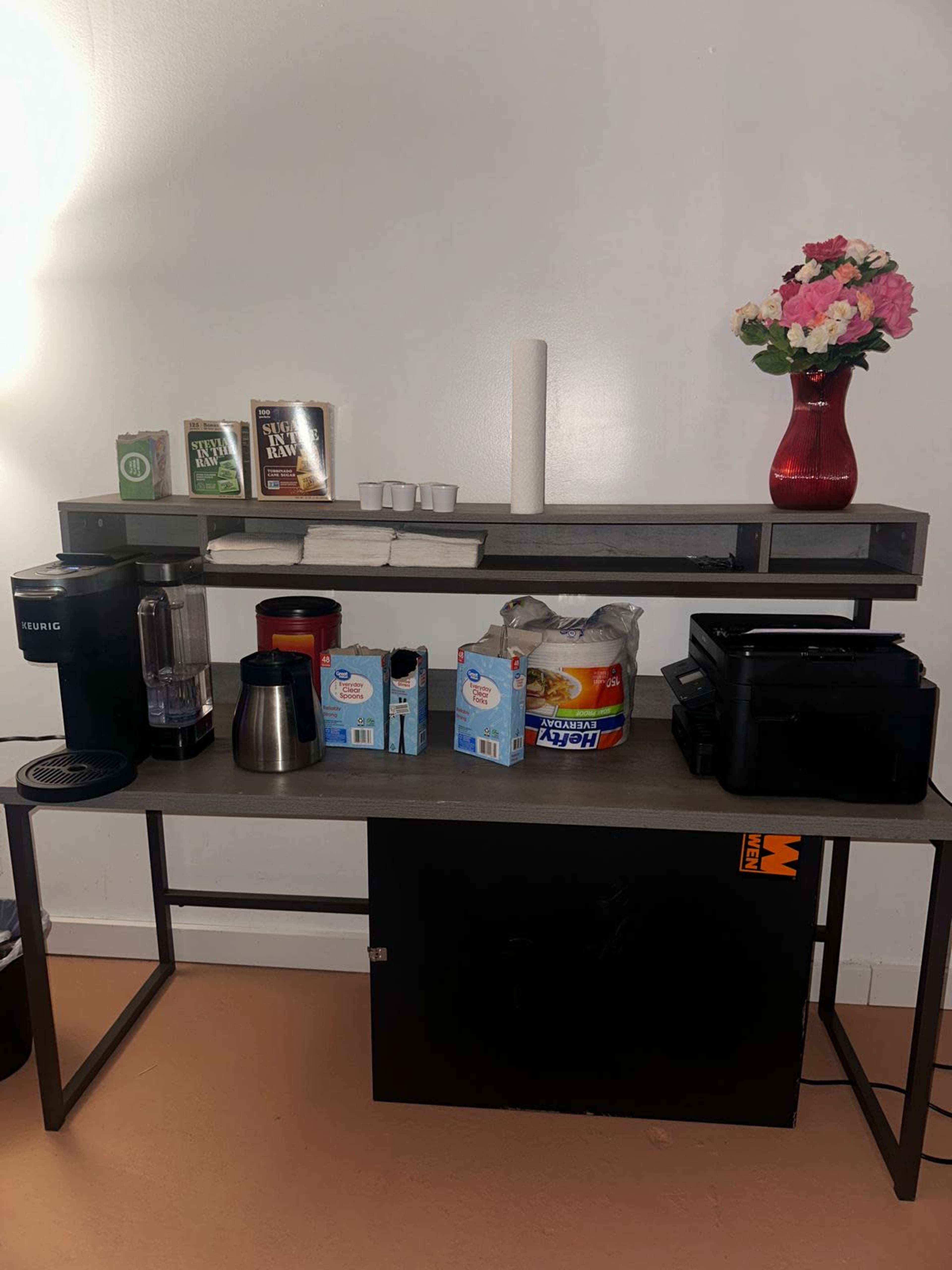 The image shows a coffee station with a Keurig coffee maker, a blender, various coffee supplies, creamers, and a vase of flowers on top of a gray shelf.