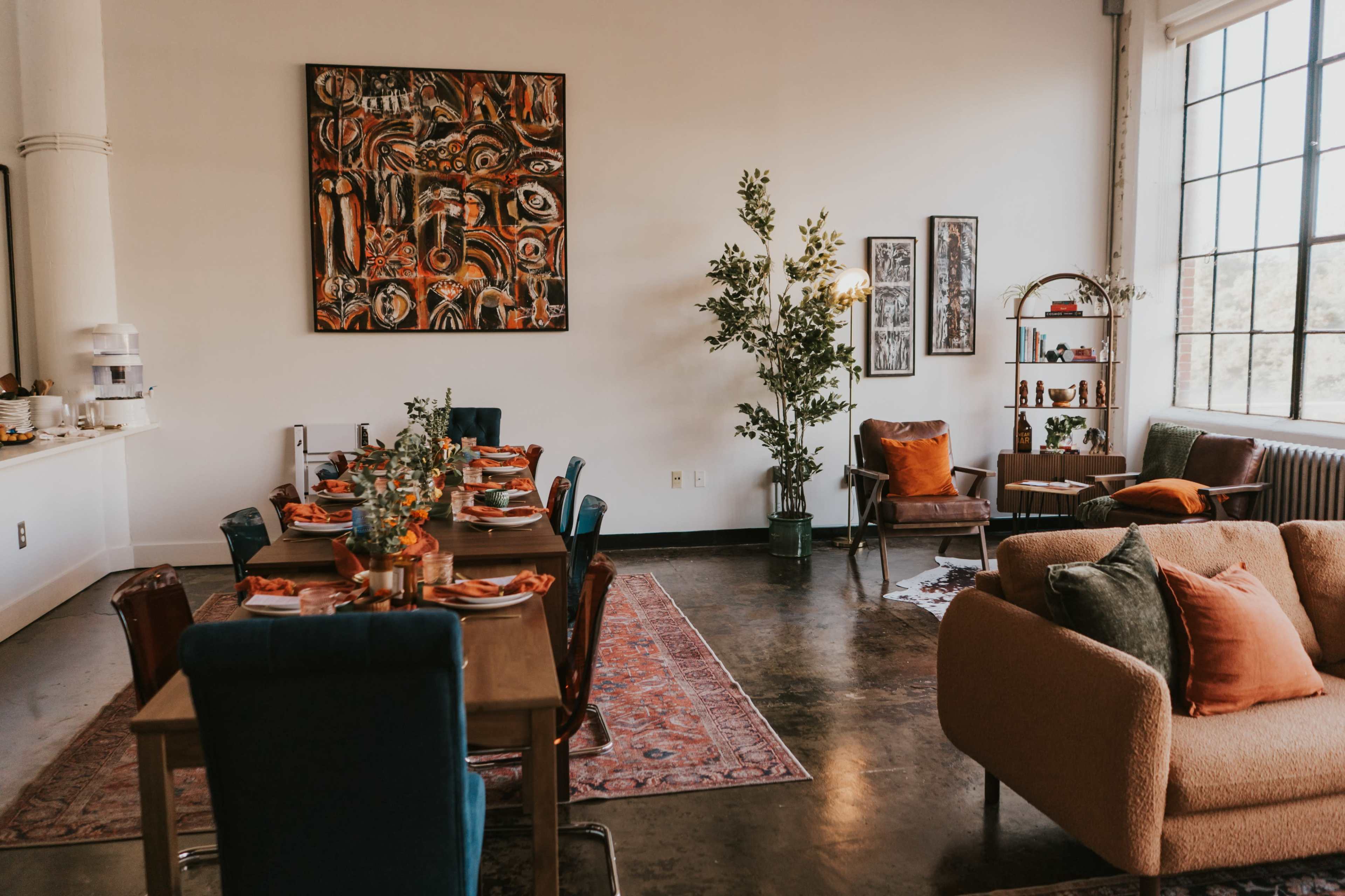 The image shows a stylish dining area with a large wooden table set for a meal, surrounded by colorful chairs, and a cozy seating arrangement in a well-lit, modern space decorated with plants and artwork.