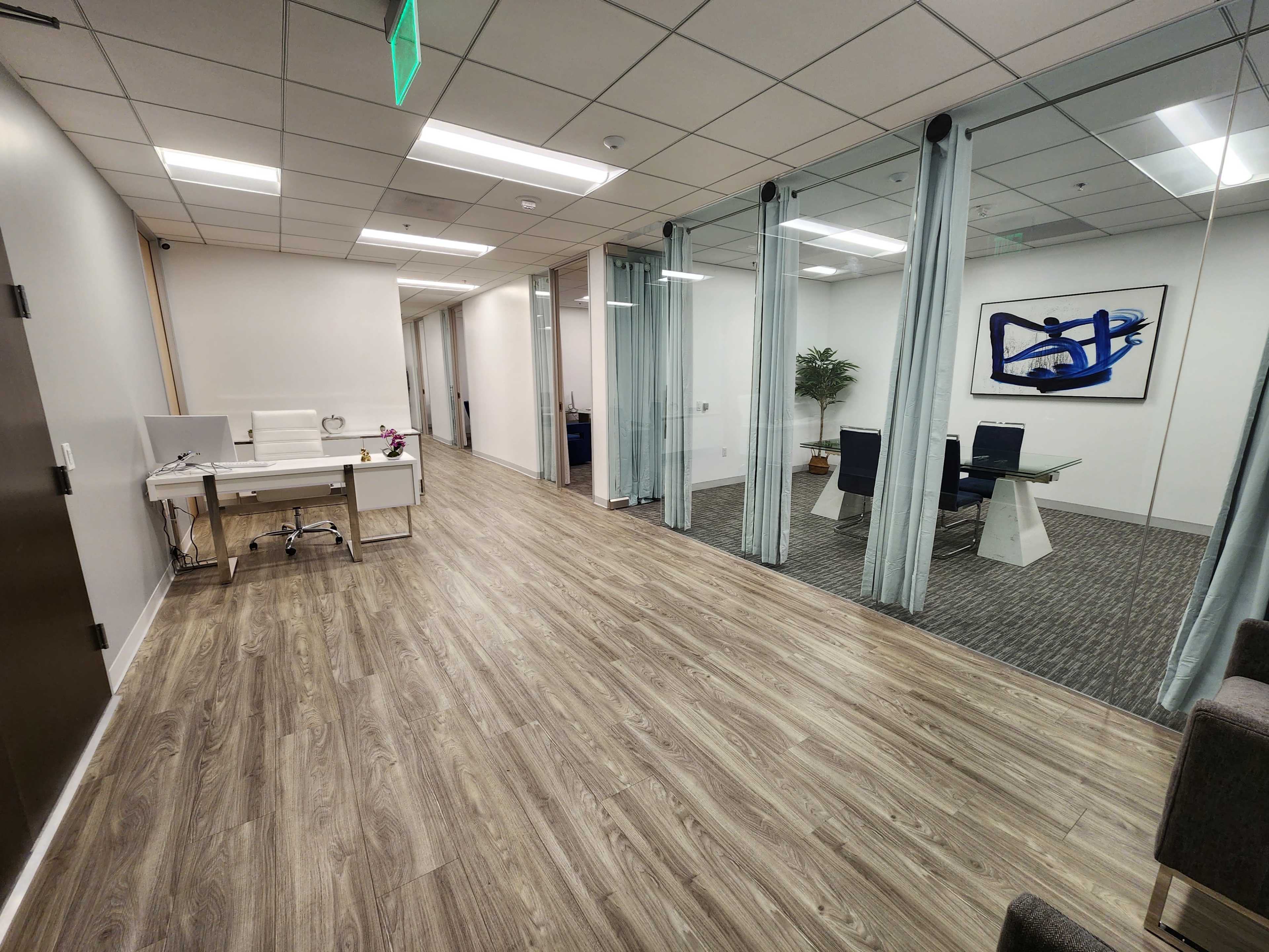 The image shows a modern office space featuring a wooden floor, a desk on the left, and glass-walled meeting rooms with a table and chairs.