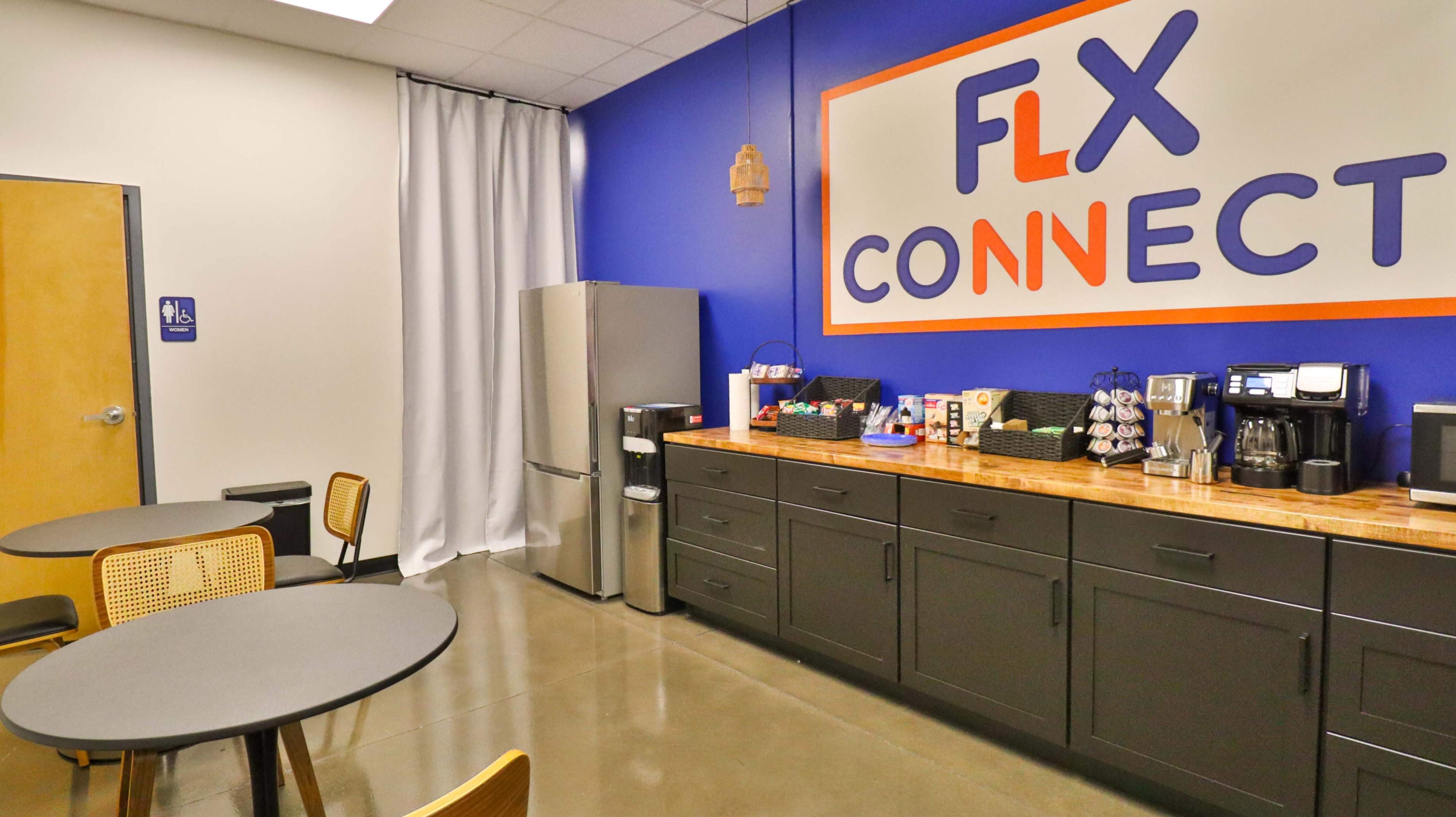 The image shows a modern break room with a snack and beverage area featuring a refrigerator, coffee machines, and seating.