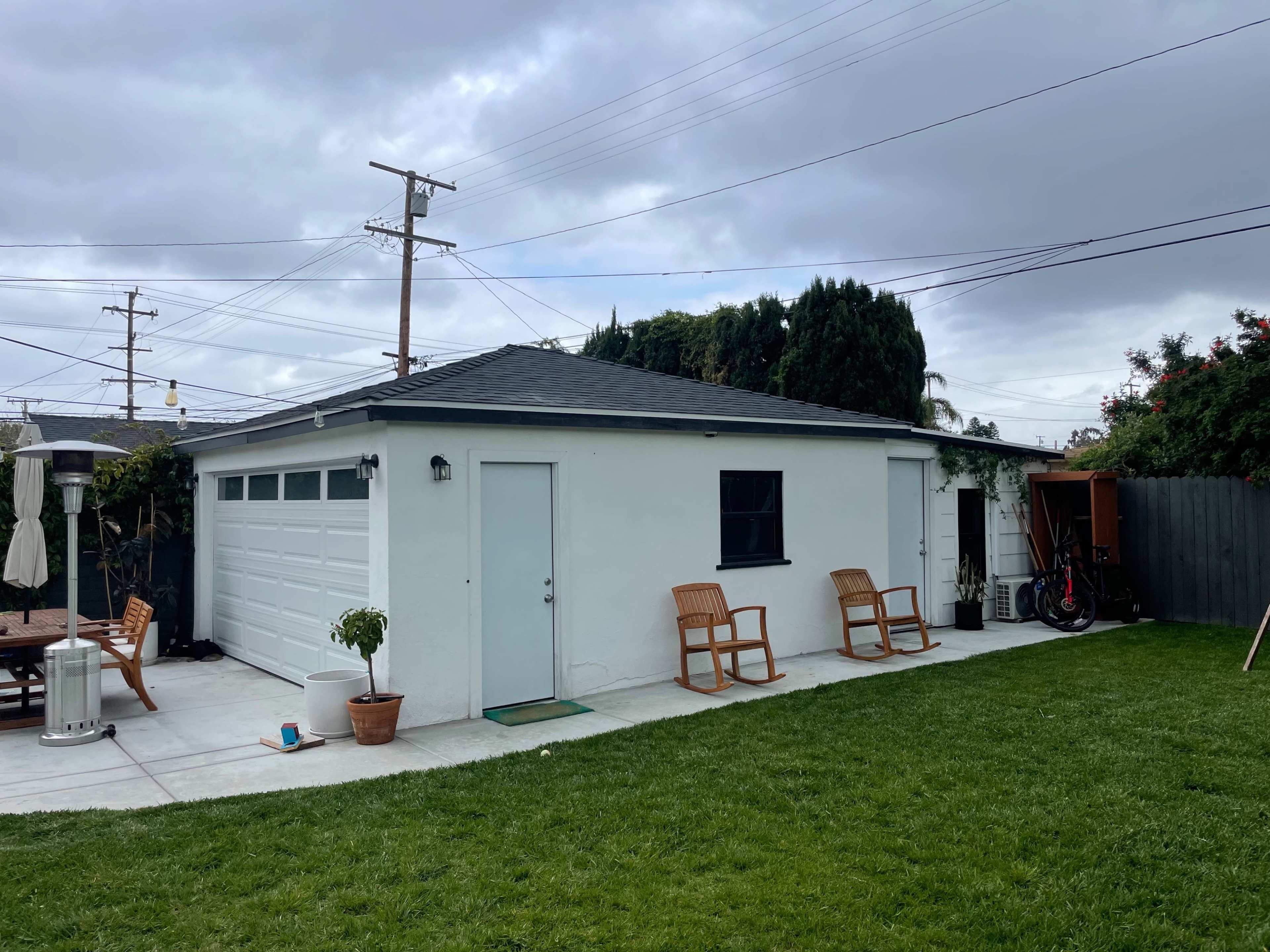 The image shows a white garage with a black roof, situated in a grassy backyard that features a patio area with chairs and a nearby shed.