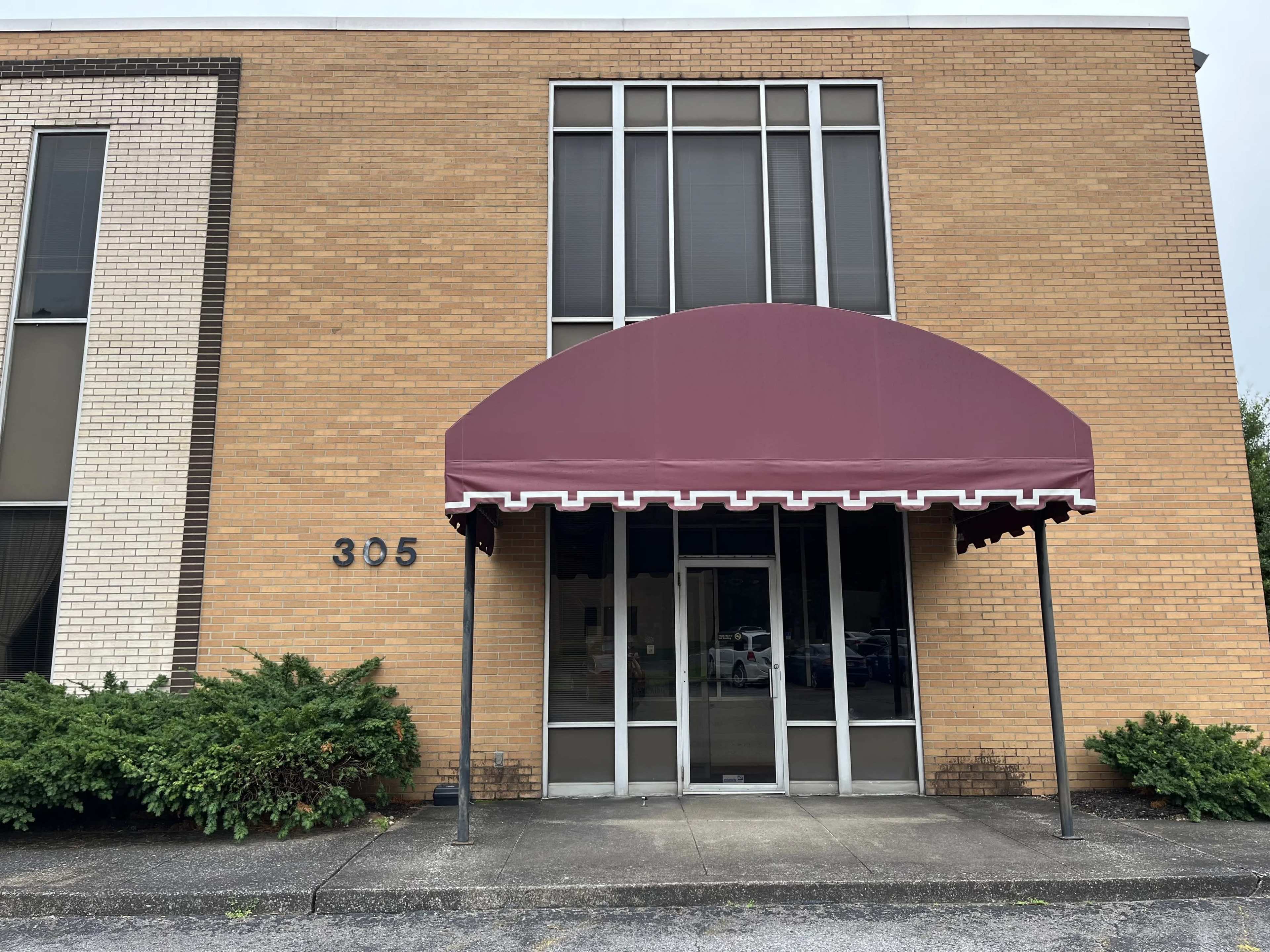 The image shows the front entrance of a brick building featuring large windows, an awning, and the number "305" displayed on the wall.