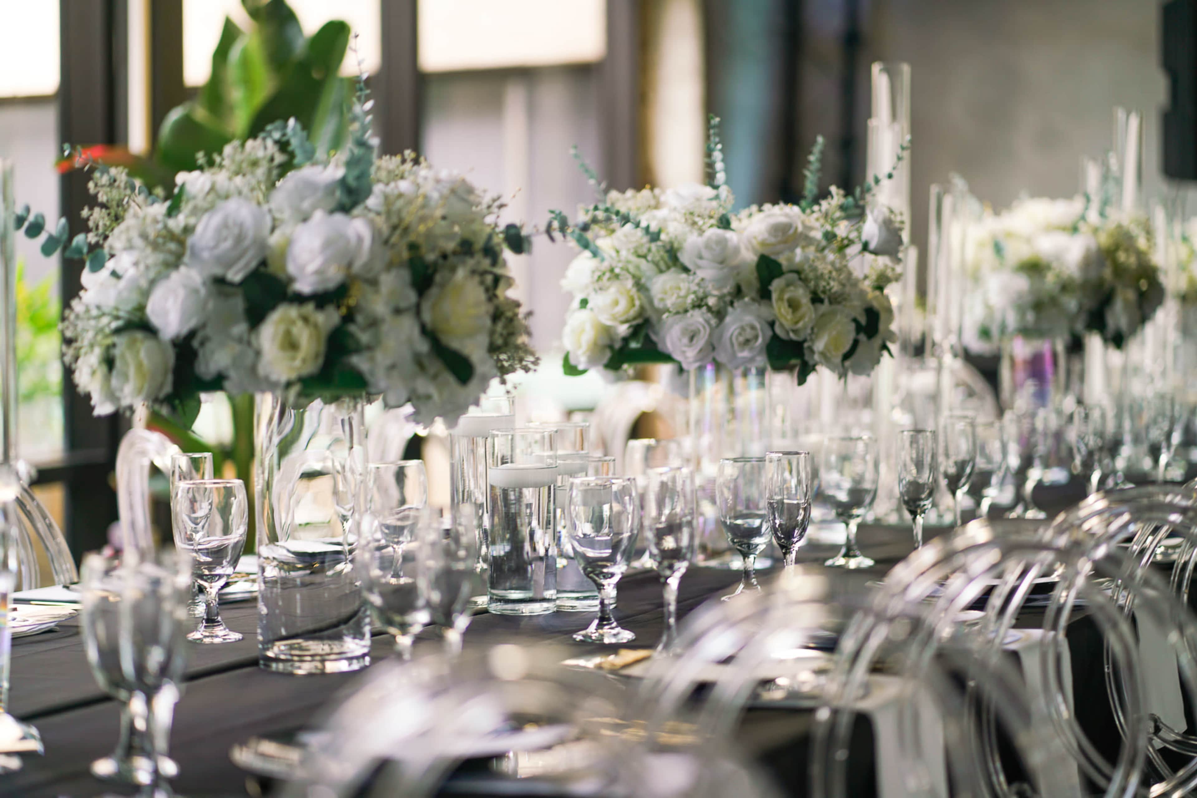 A long dining table is set with clear glassware and elegant floral centerpieces featuring white roses and greenery.