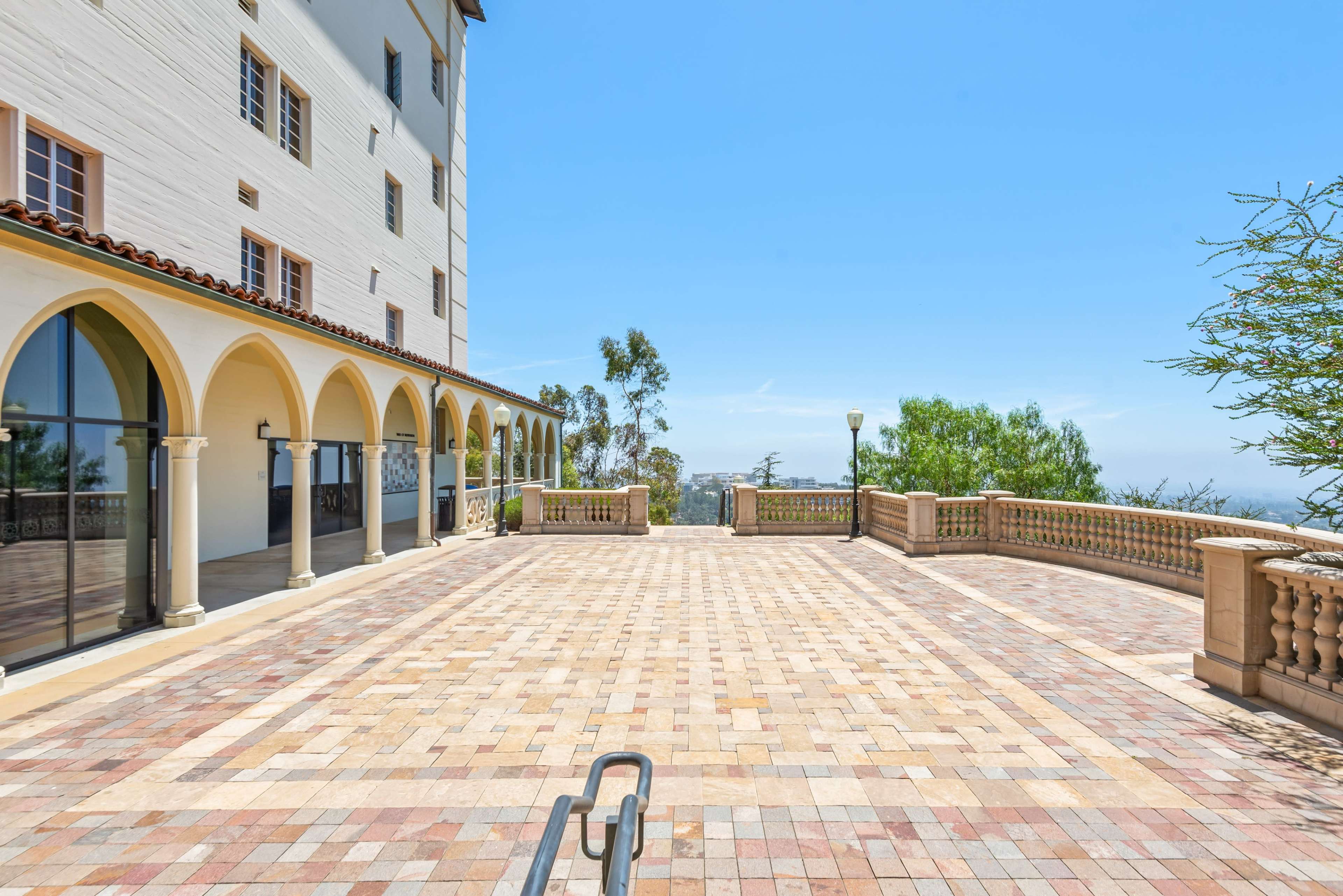 The image shows a spacious outdoor terrace with a tiled surface and arched columns, adjacent to a large building under a clear blue sky.