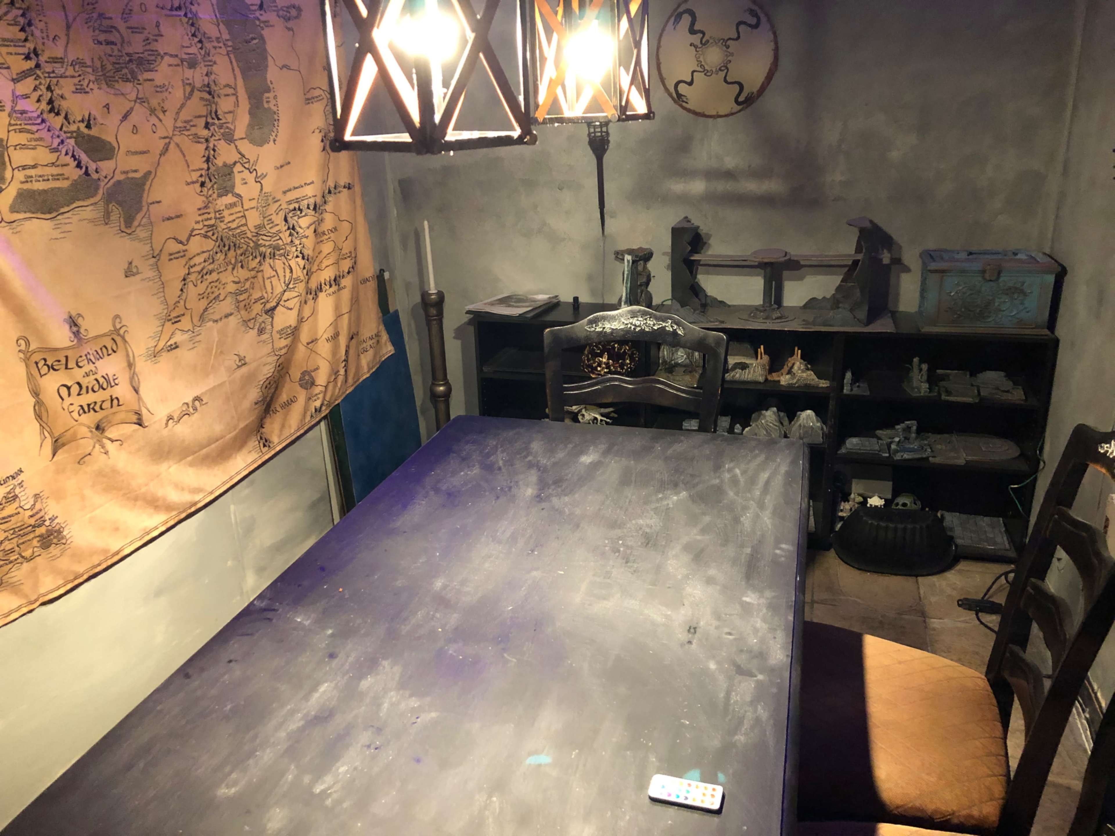 The image shows a dimly lit room featuring a large wooden table surrounded by chairs, with a map hanging on the wall and shelves displaying various items.