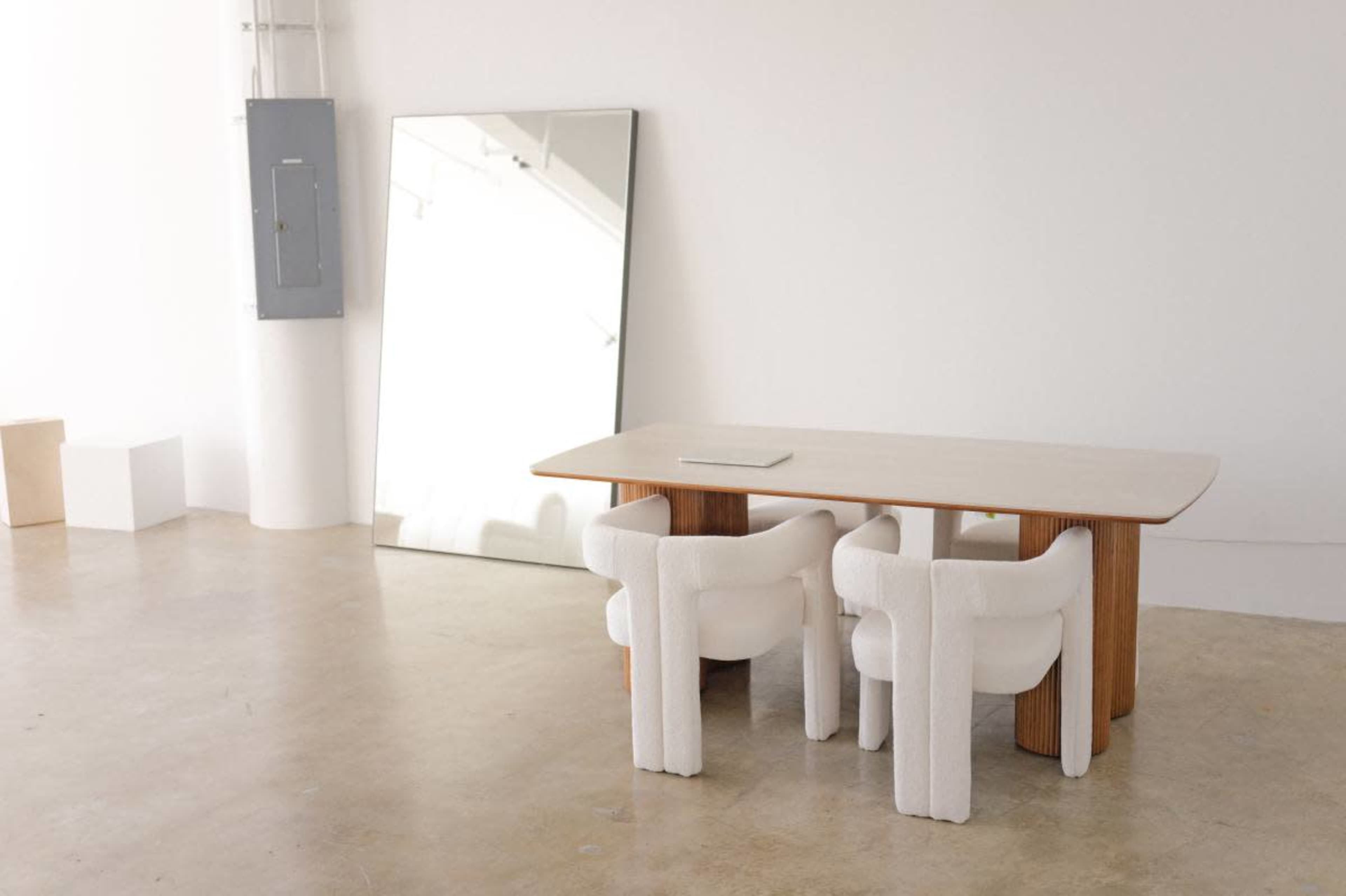 A minimalist room features a wooden table with three upholstered chairs and a large mirror against a white wall.