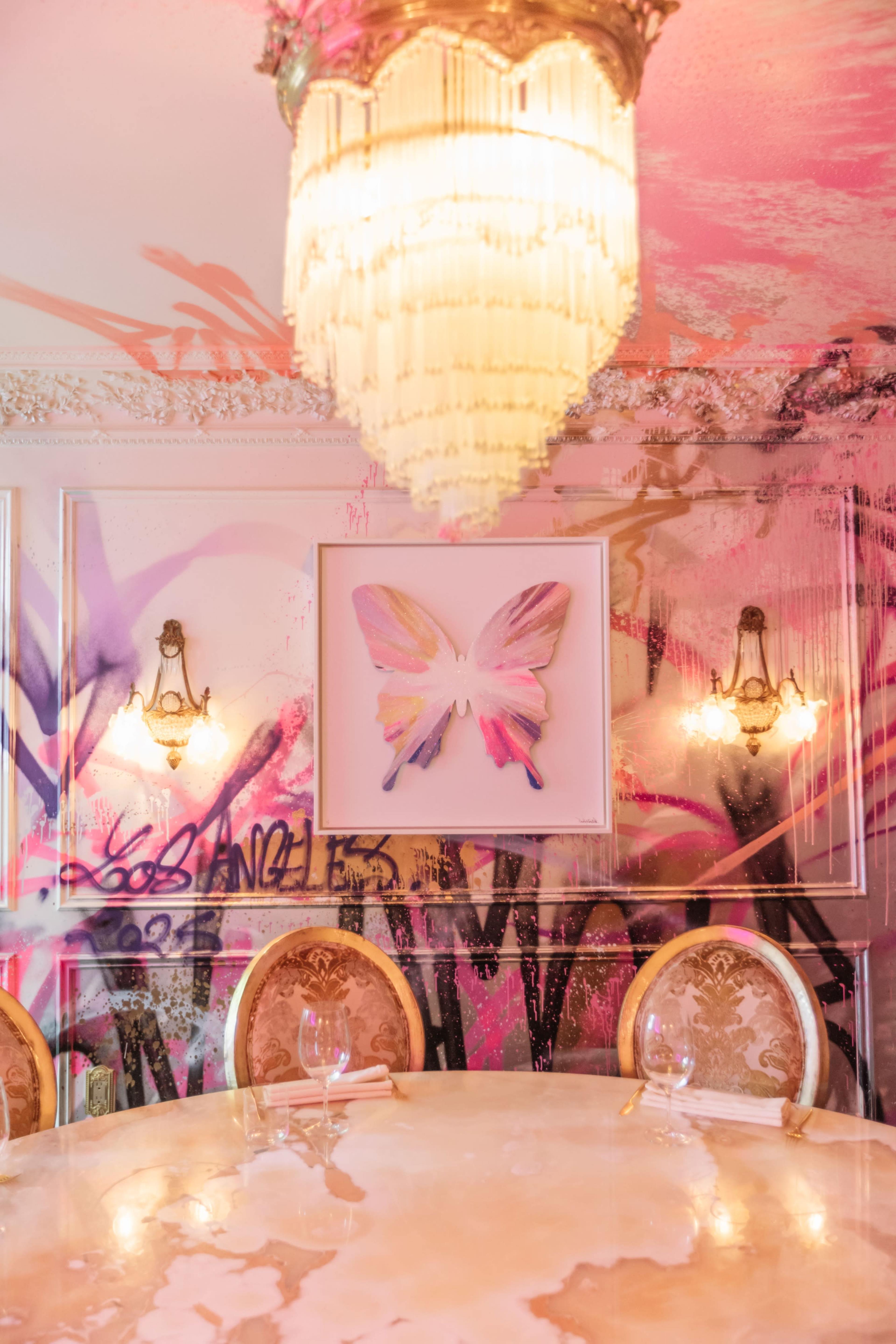 A round marble table is set with glassware under a chandelier, surrounded by ornate chairs, with a colorful butterfly artwork and graffiti on the walls.