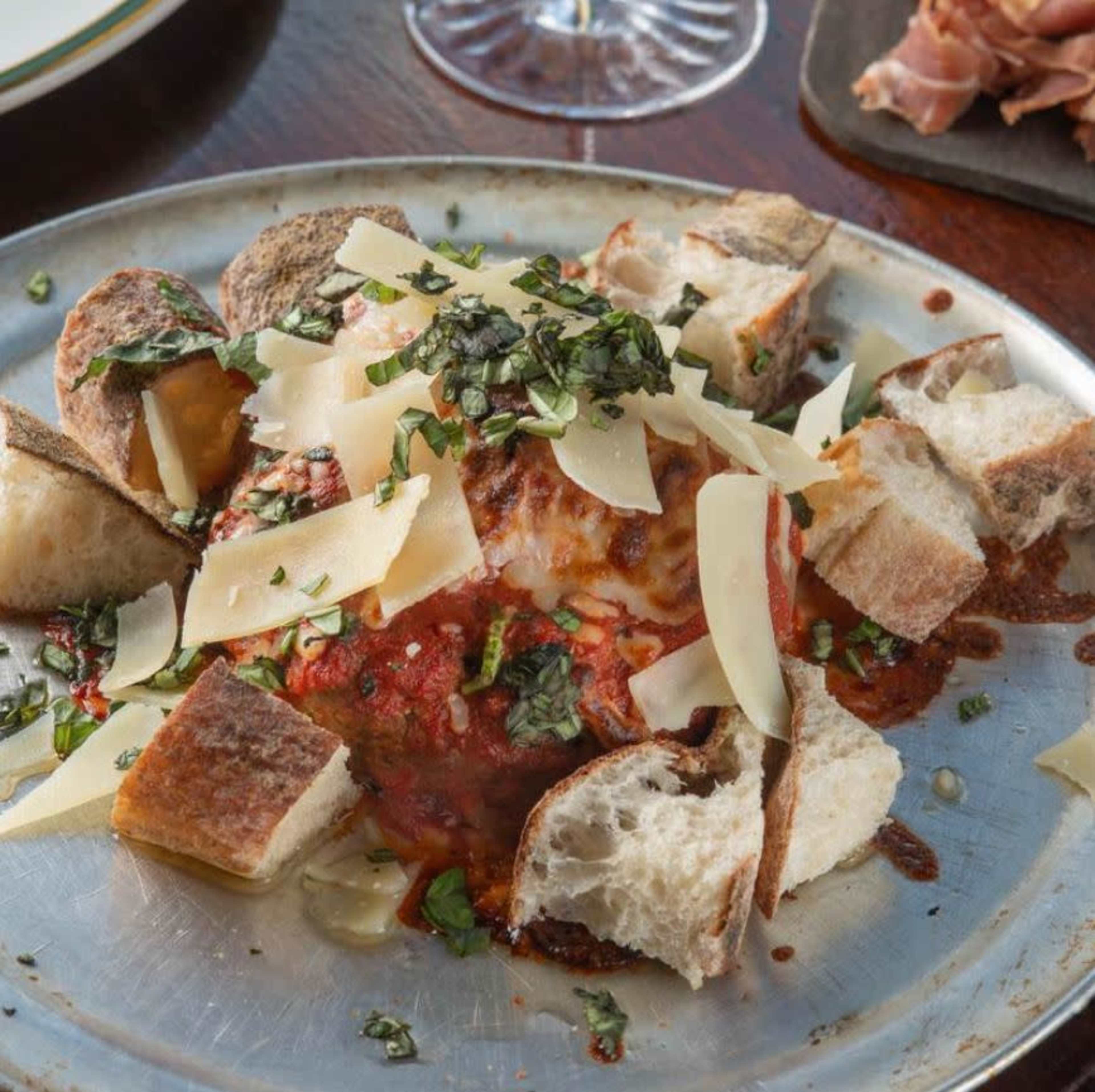The image shows a large meatball topped with melted cheese, garnished with fresh herbs, and surrounded by pieces of bread on a metal platter.