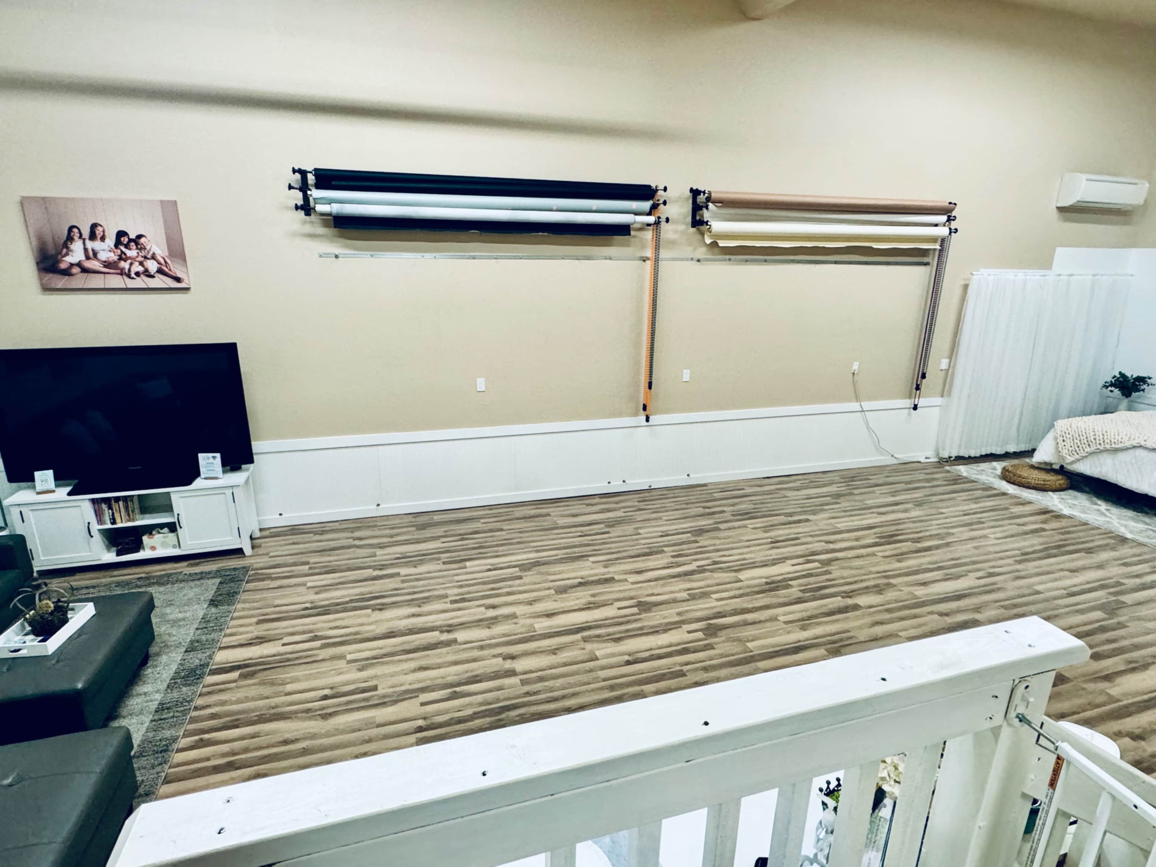 The image shows a spacious studio with wooden flooring, equipped with a television, a backdrop system mounted on the wall, and a cozy area with a bed and plants.