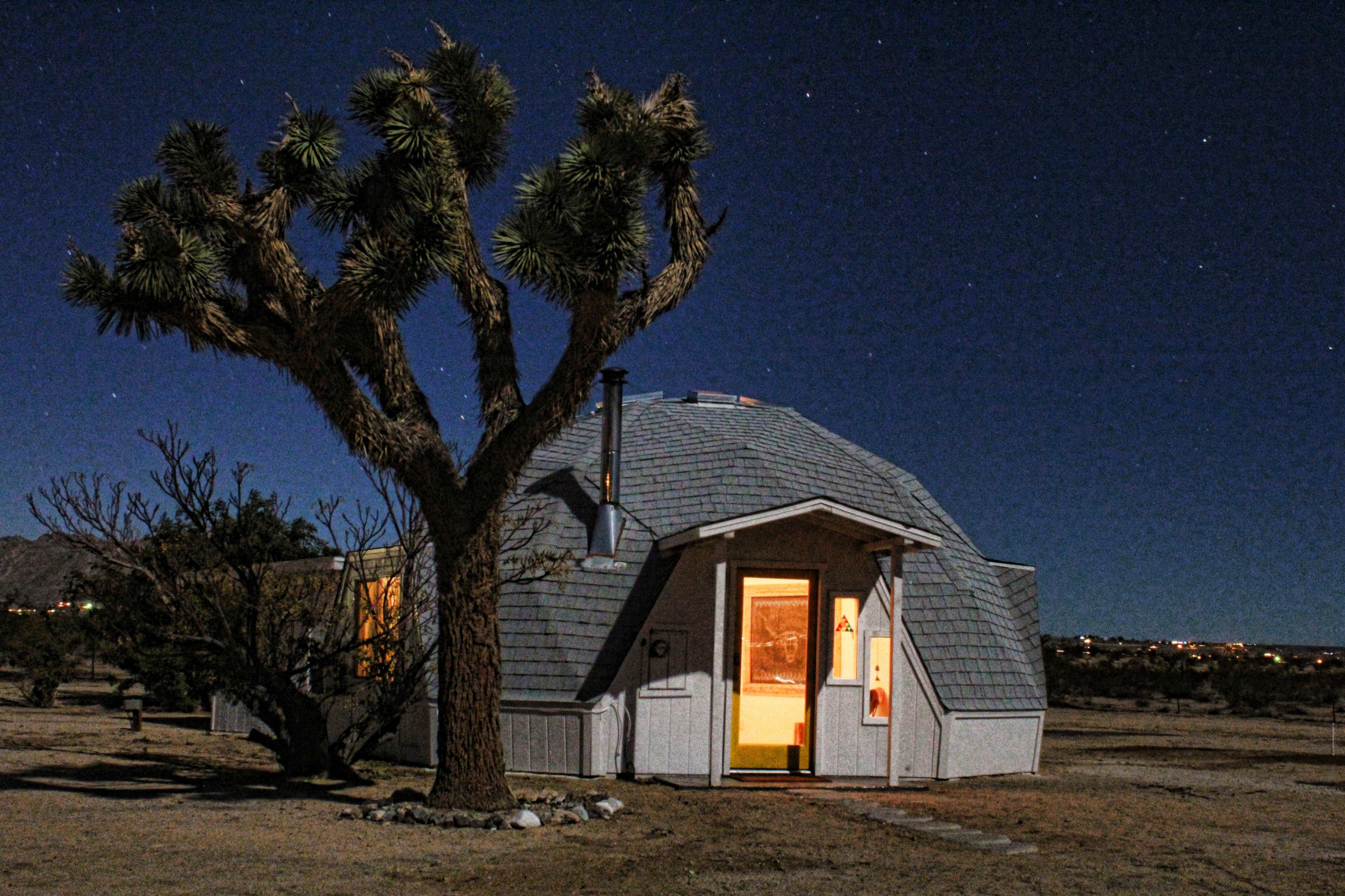 A geodesic dome house is illuminated at night, surrounded by desert vegetation under a starry sky.