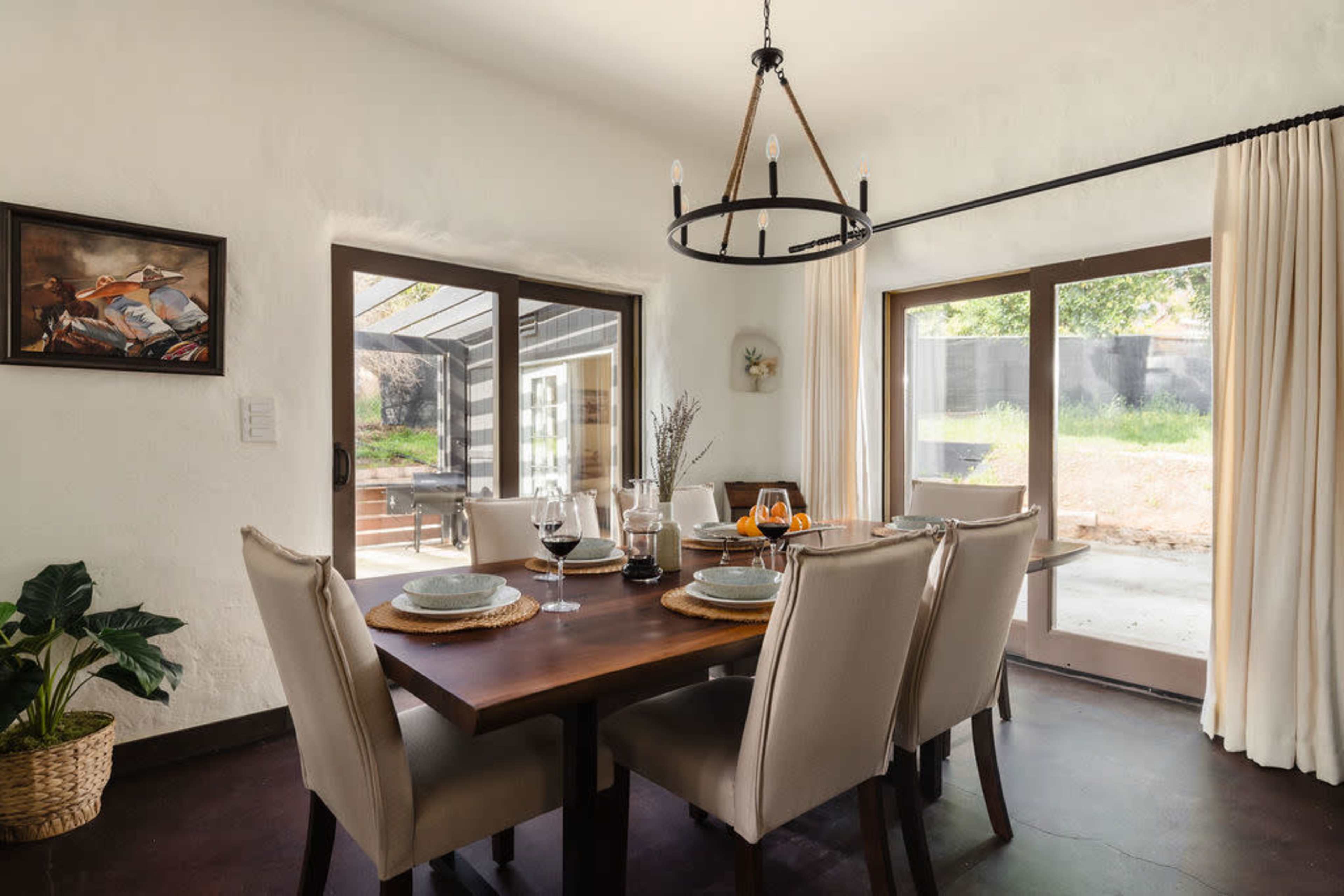A dining room features a wooden table set for six, surrounded by upholstered chairs, with large windows providing views of an outdoor patio.