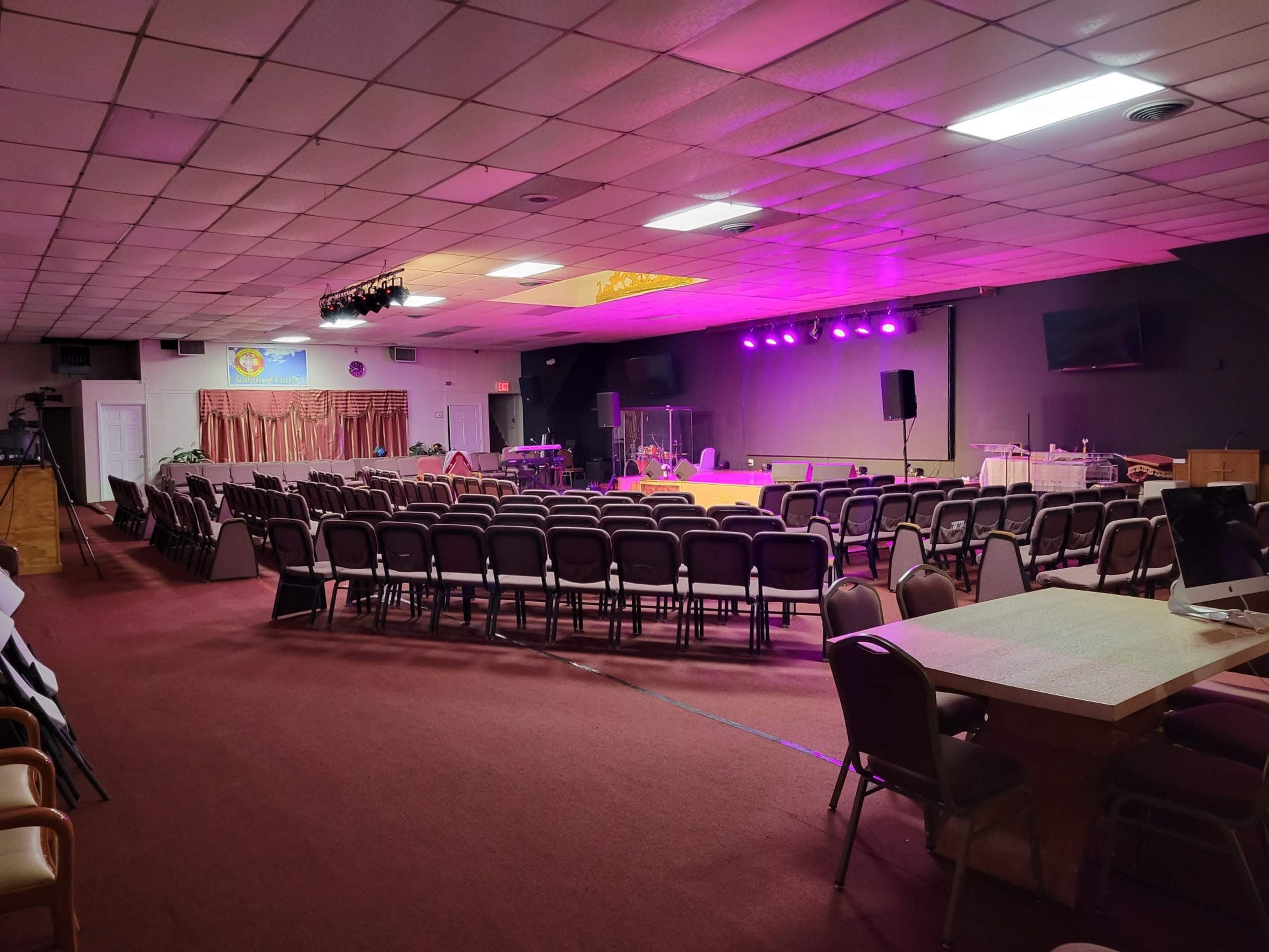 A spacious auditorium features rows of chairs facing a stage with purple lighting, flanked by sound equipment and a small band setup.