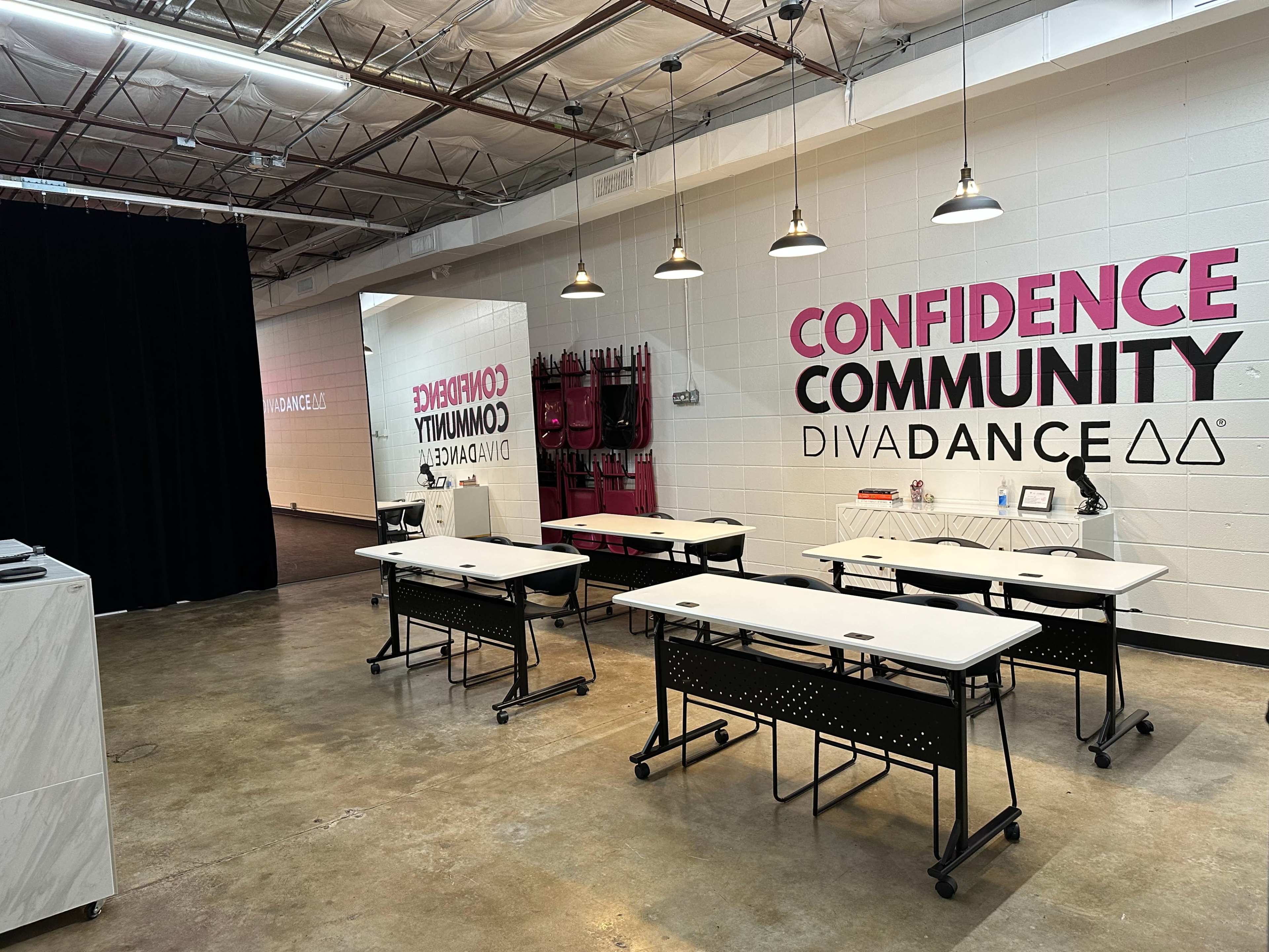 A spacious room with several tables arranged for a class or event, featuring a wall with the words "CONFIDENCE," "COMMUNITY," and "DIVA DANCE" in bold lettering.