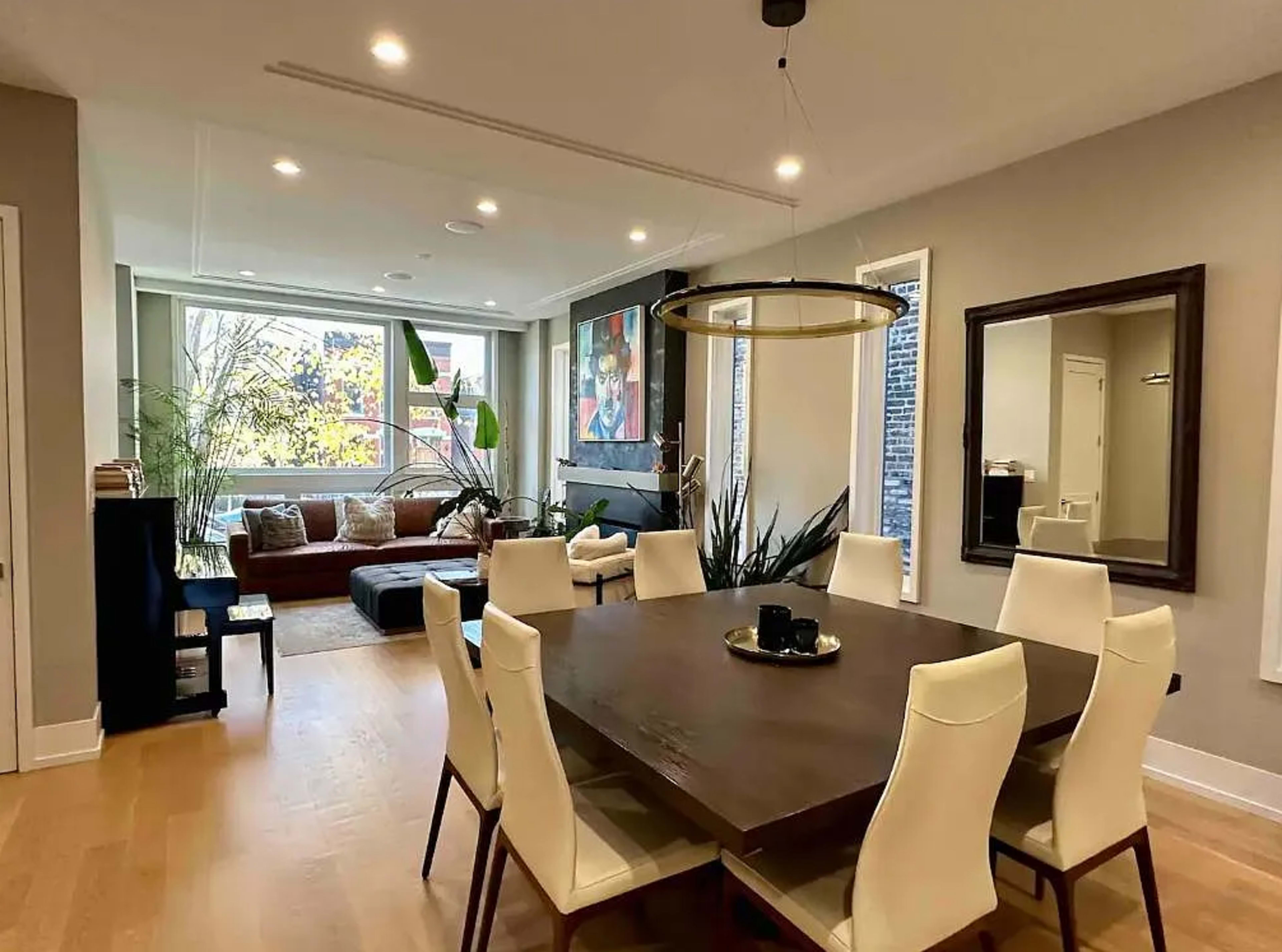 A modern dining room with a wooden table surrounded by white chairs leads into a living area featuring a sofa and large windows with plants and artwork on the walls.