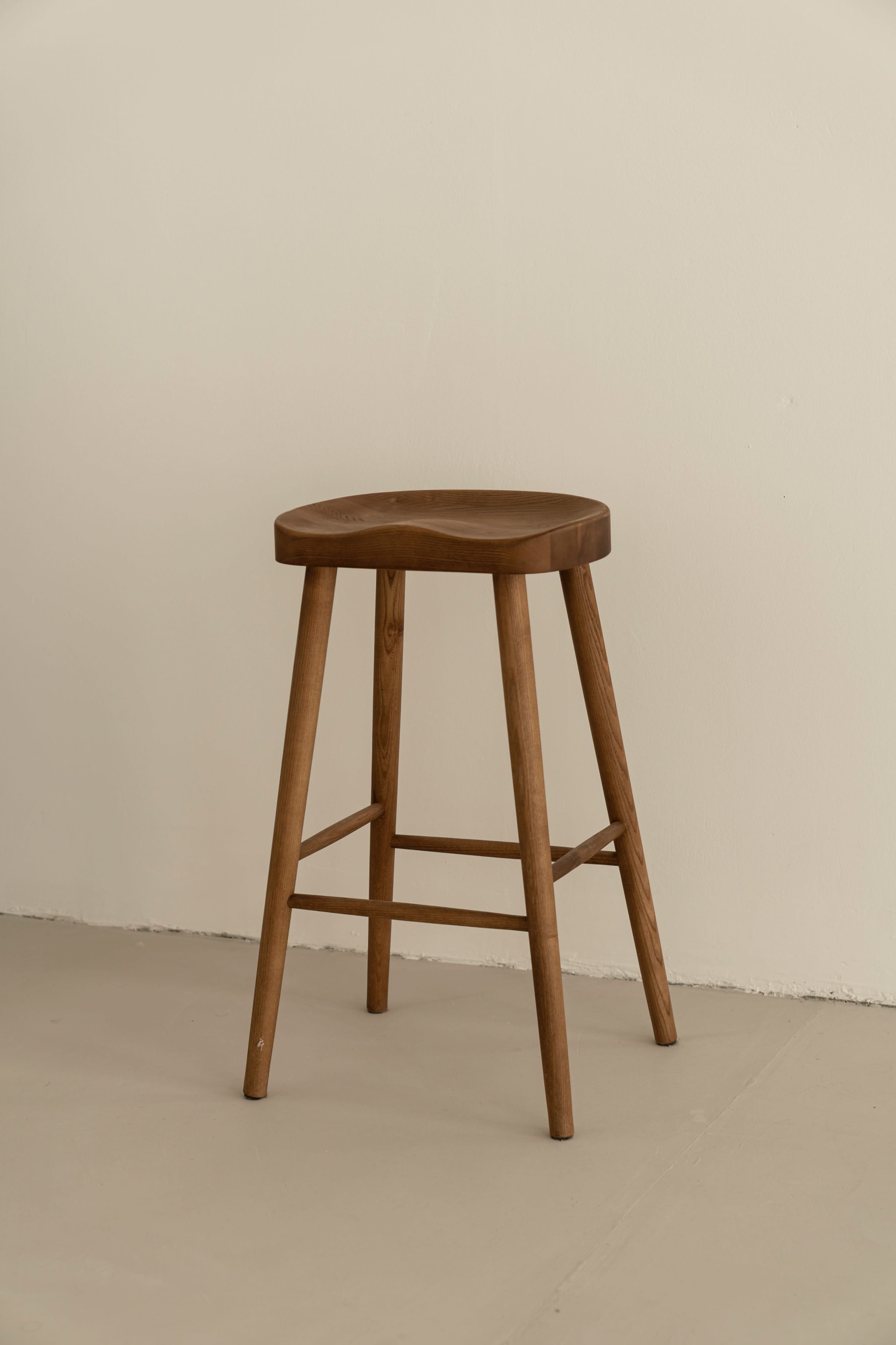 A wooden stool with three legs stands against a plain wall.