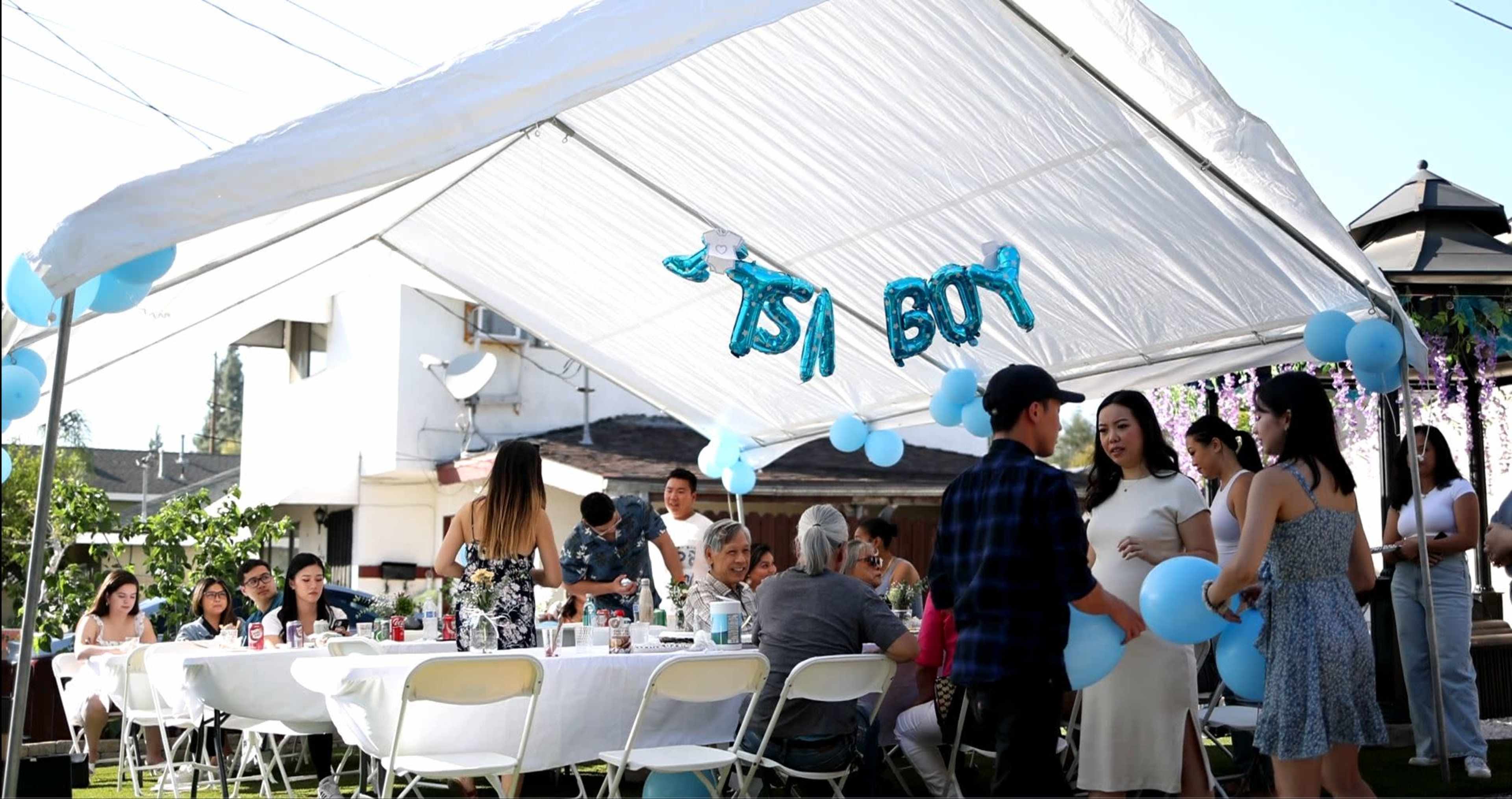 A large outdoor gathering under a tent features tables set with food and decorations, including a "1st BDAY" balloon banner and guests socializing.
