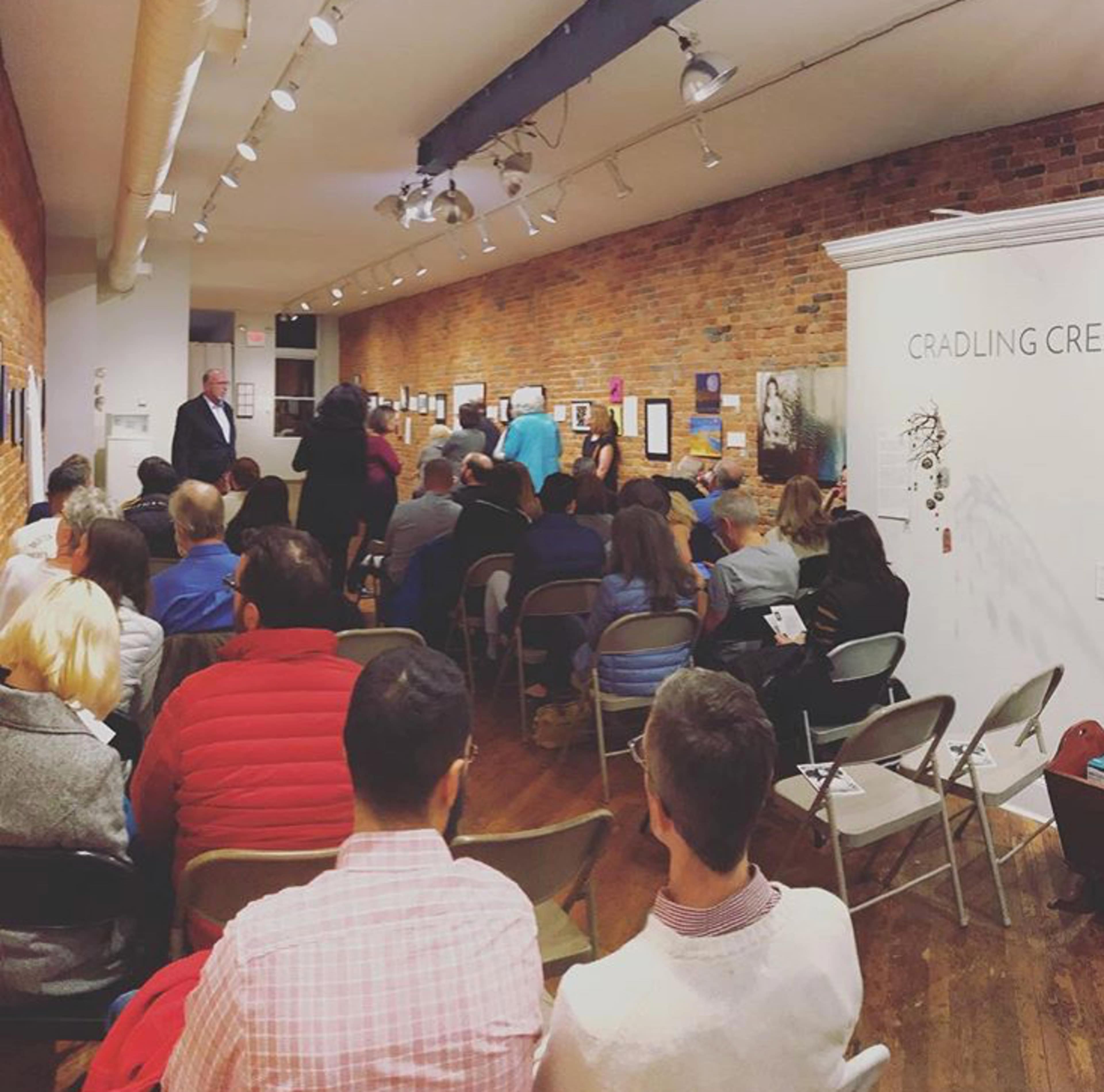An audience seated in rows for an event in an art gallery, with a display of artworks and a brick wall in the background.