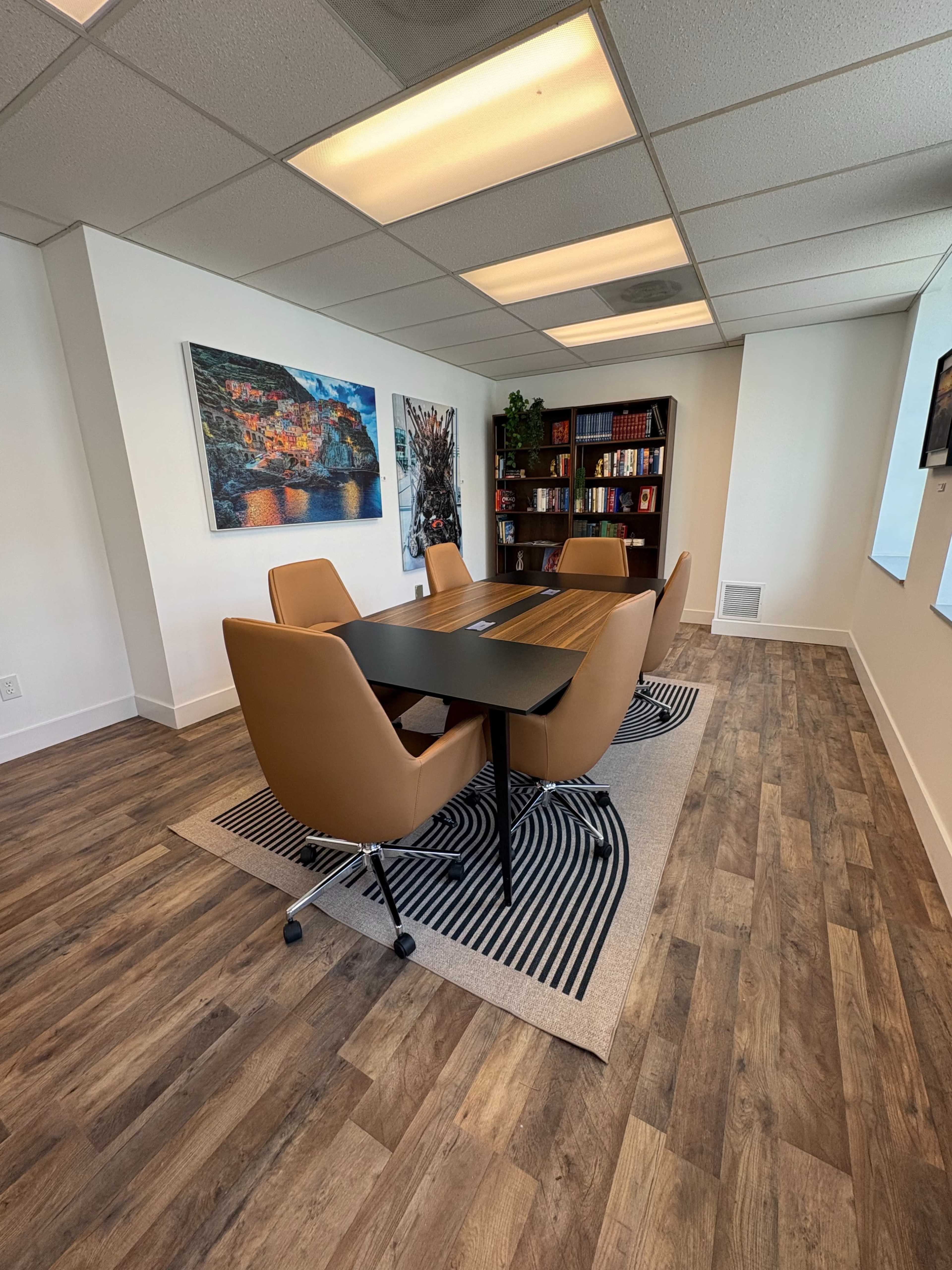 A conference room features a dark table surrounded by four brown chairs, a rug on the floor, a bookshelf filled with books, and a large artwork on the wall.