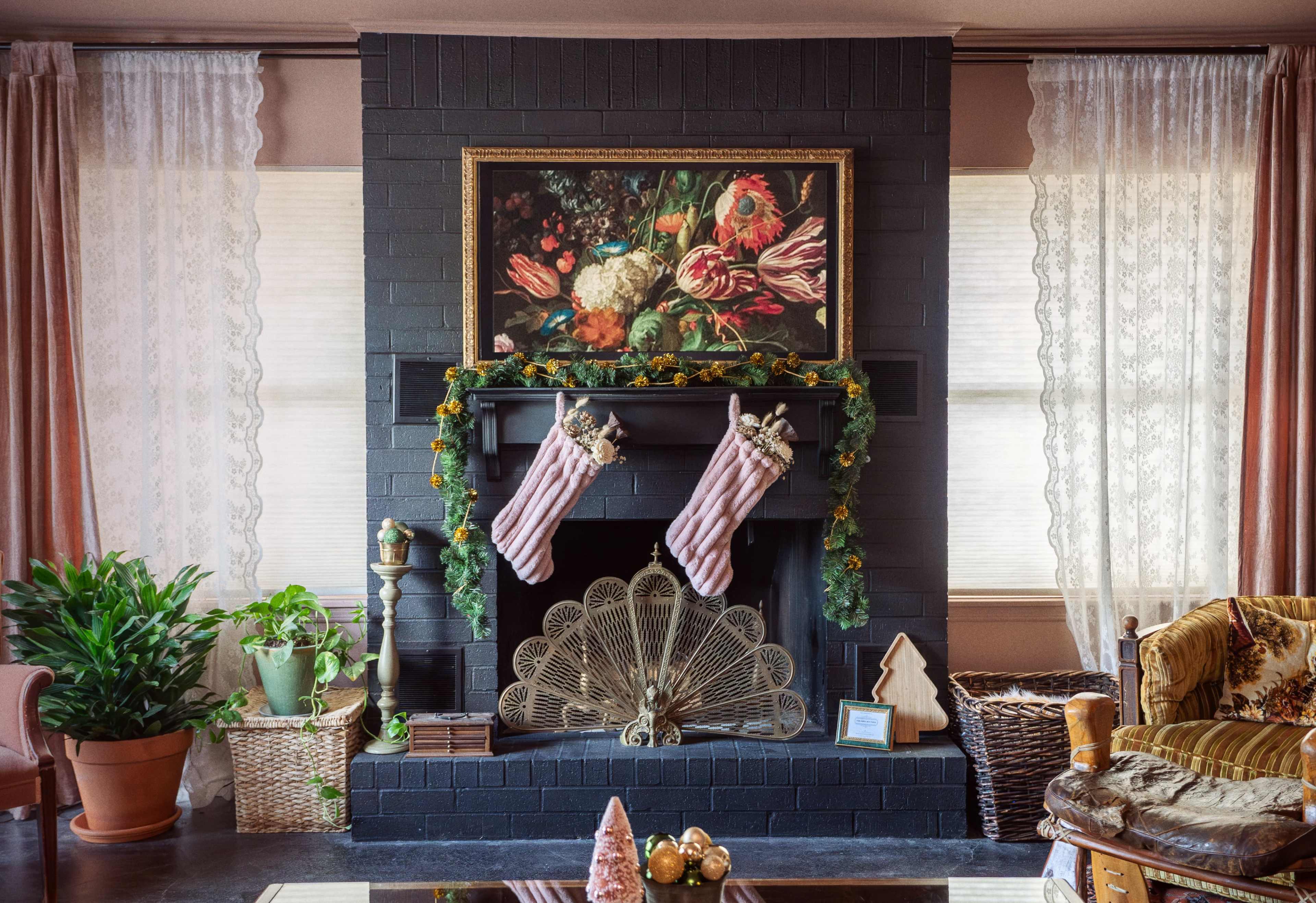 A living room with a black fireplace adorned with a floral painting, two stockings, and decorative greenery, surrounded by curtains and plants.