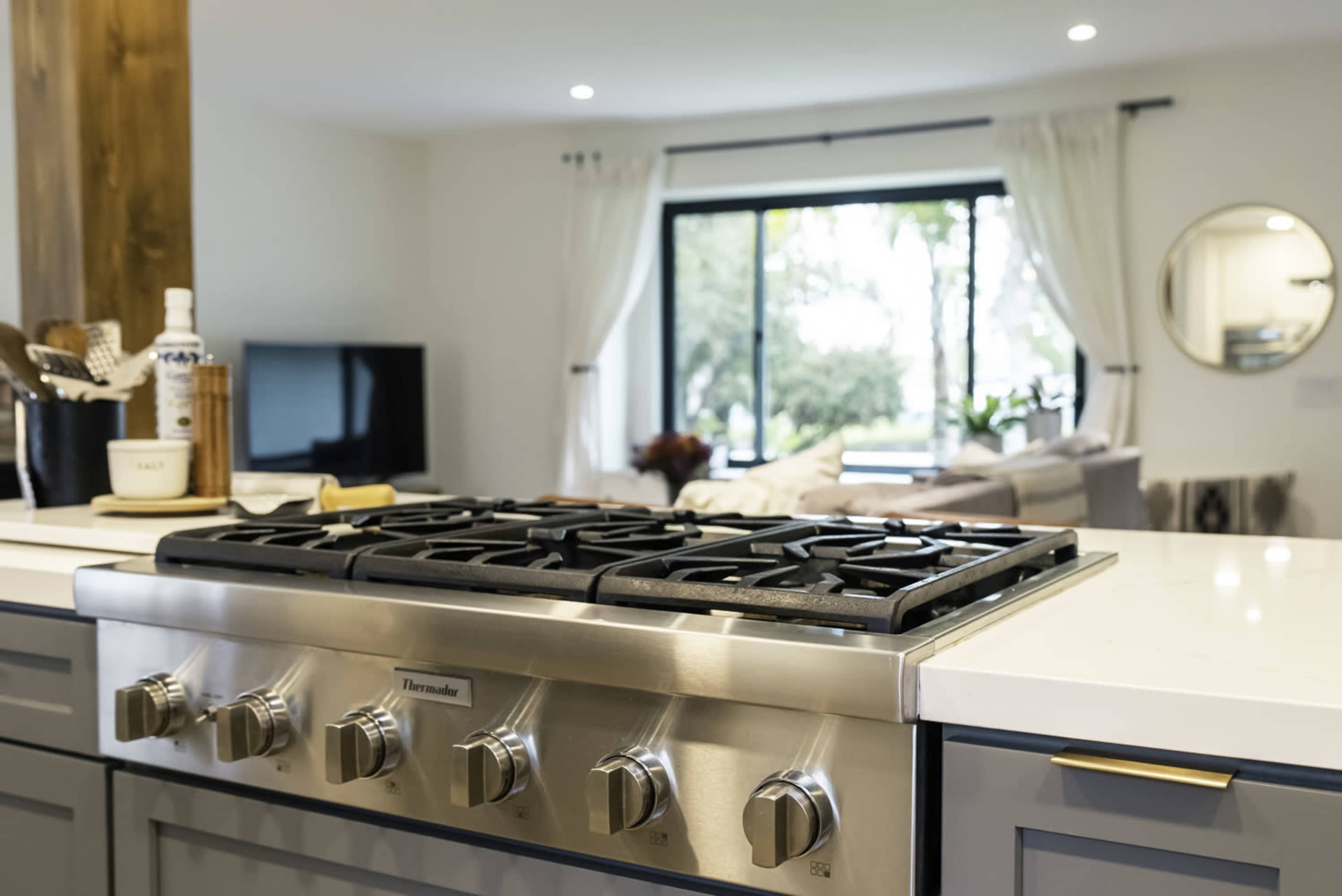 The image shows a modern kitchen countertop with a stainless steel gas stove in the foreground and a bright living area visible through a large window in the background.
