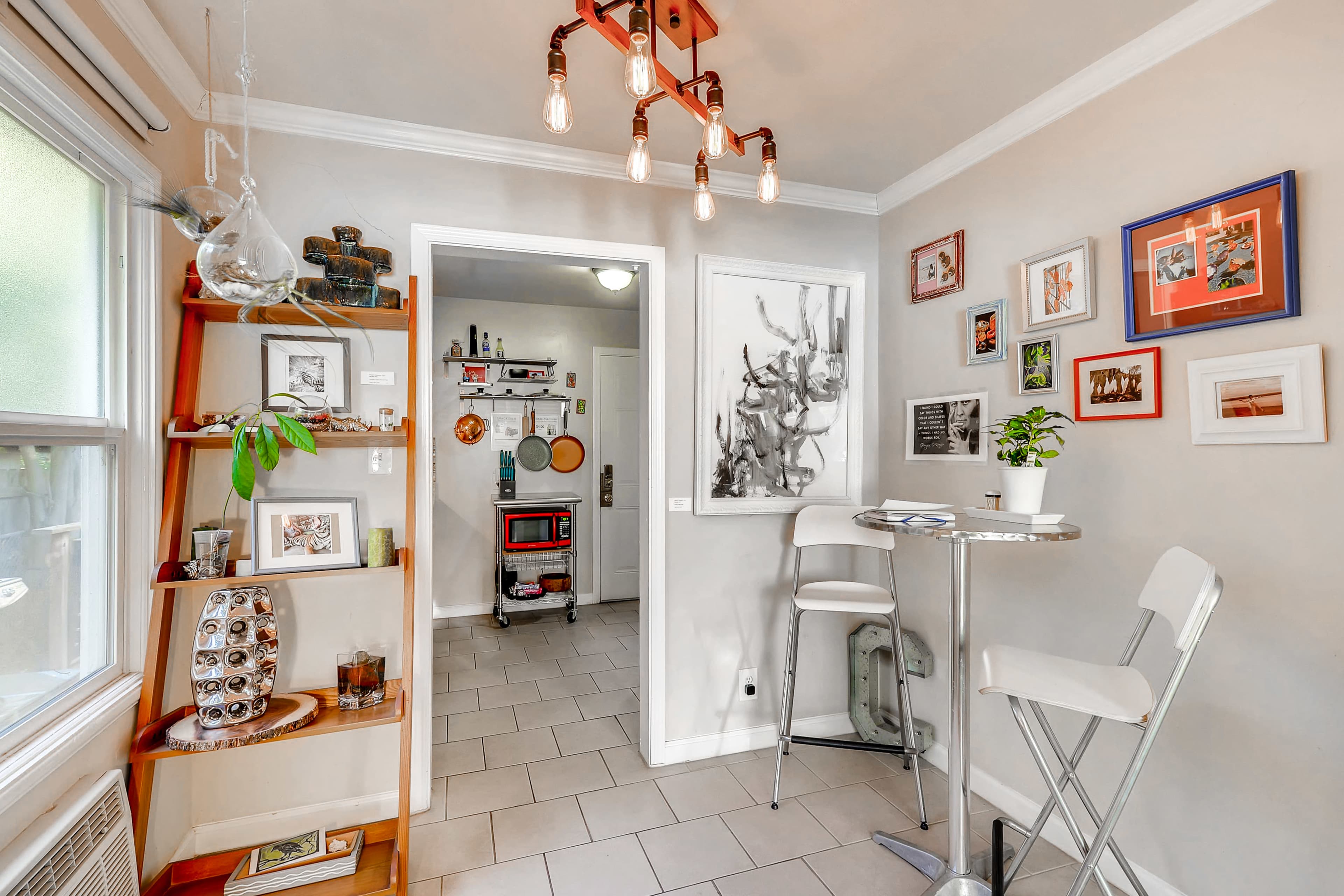 The image shows a small dining area with a round table and two white chairs, adjacent to an entryway featuring a red cabinet and various framed pictures on the wall.