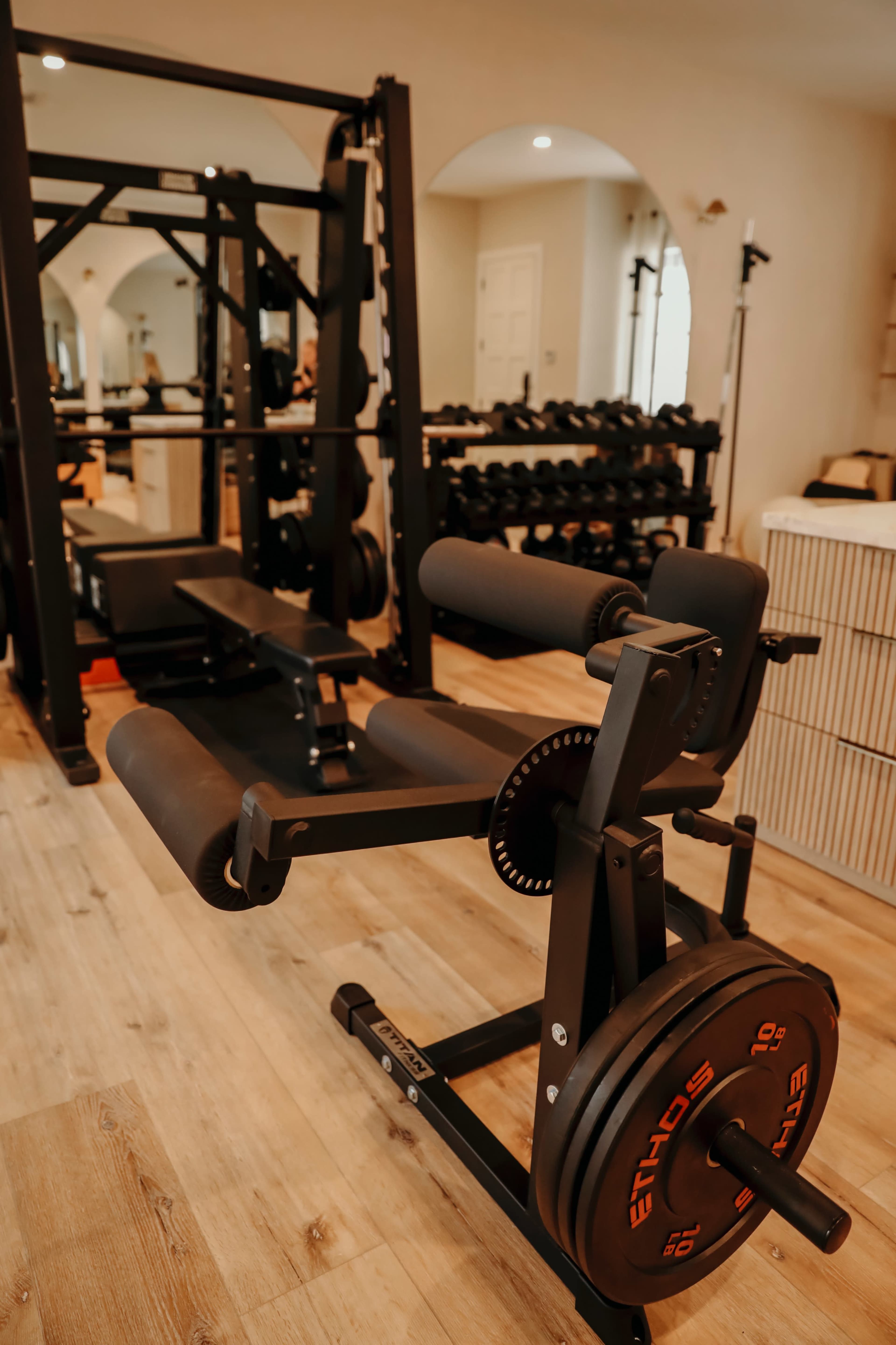 The image shows a home gym setup featuring a weight bench and a rack of dumbbells in a well-lit room with wooden flooring.