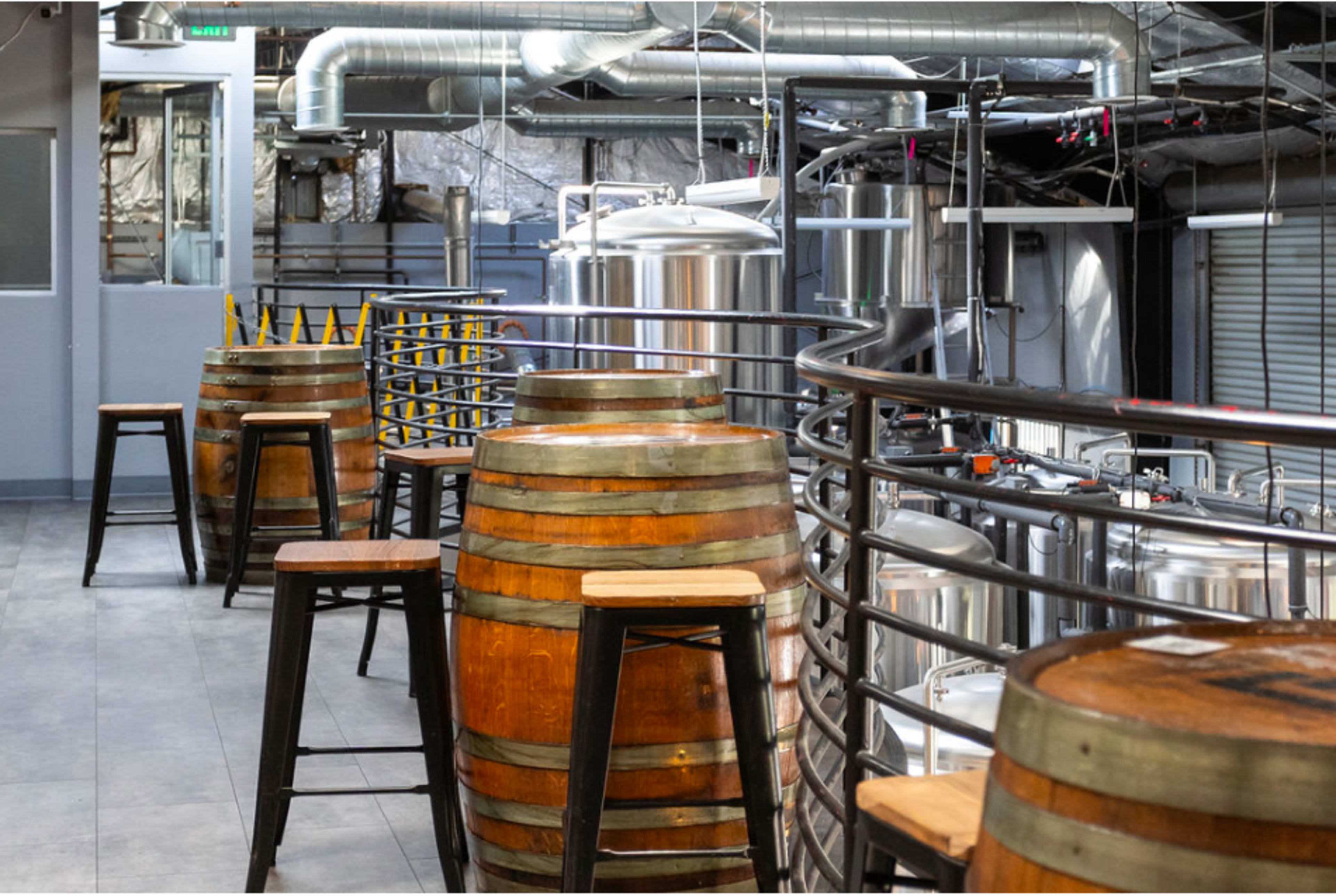 A modern brewery interior with wooden barrels and high stools arranged along a metal railing.