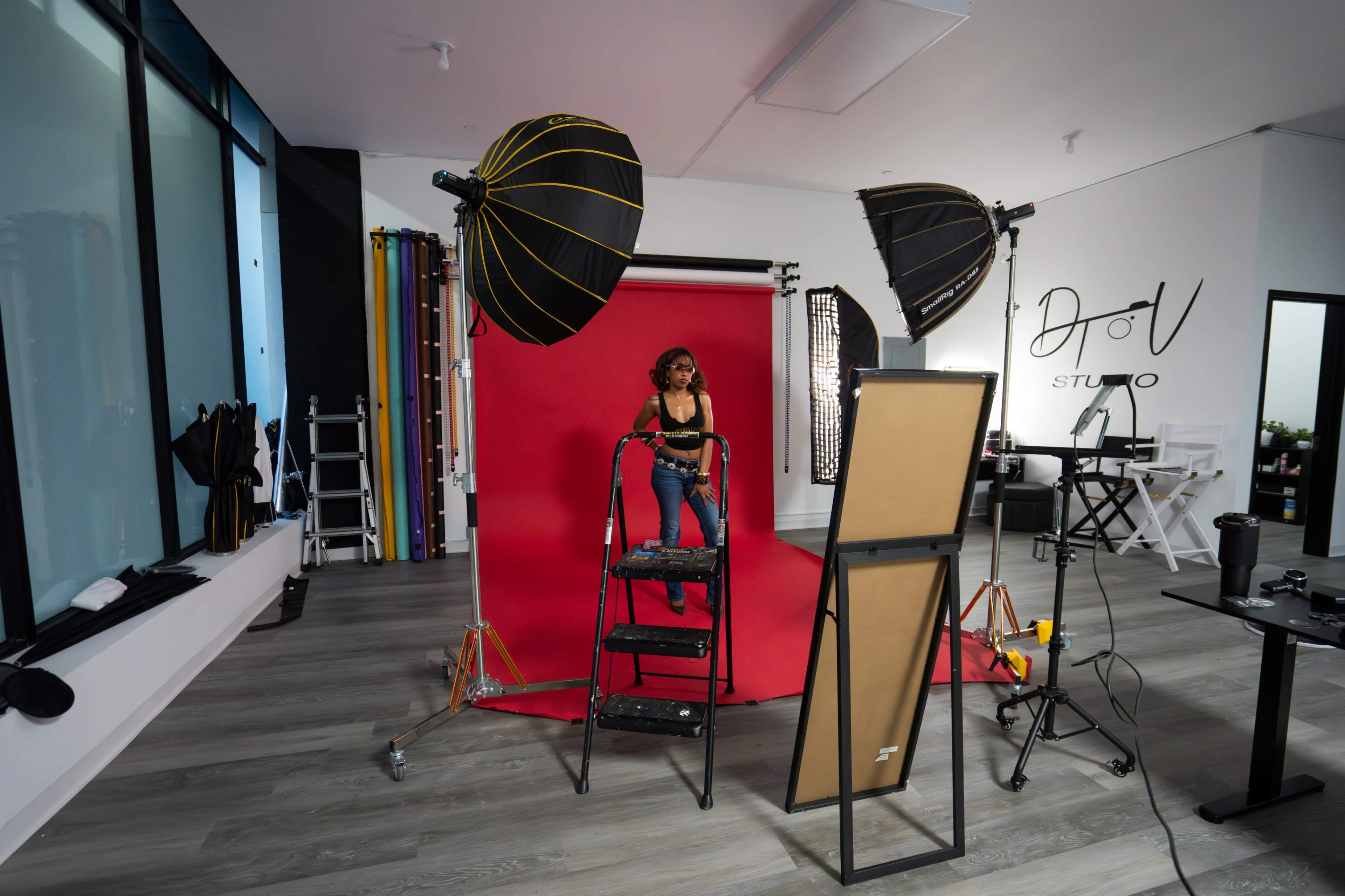 A model stands on a ladder in front of a red backdrop inside a photography studio equipped with lighting and various props.