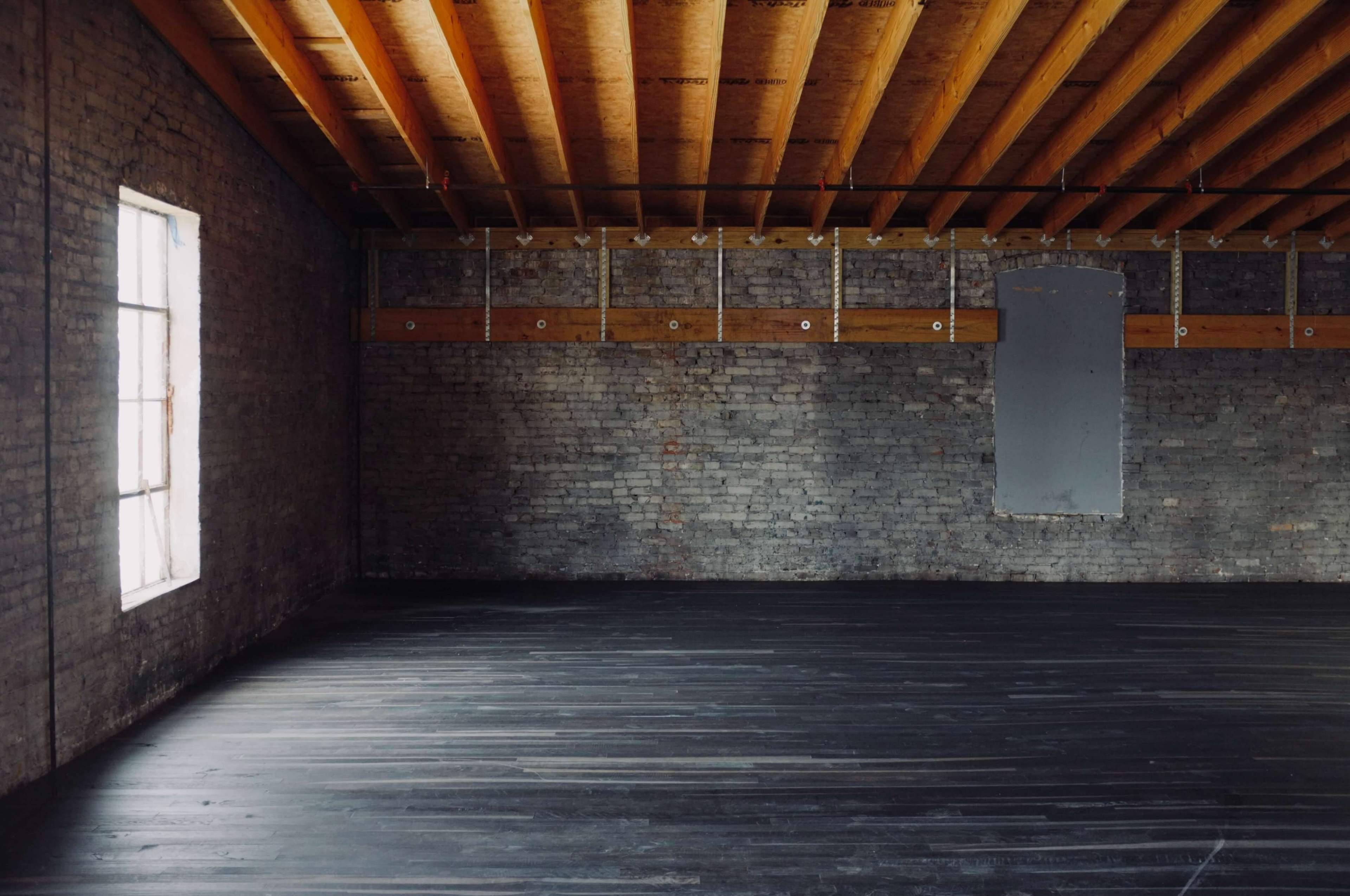 The image shows an empty room with exposed wooden beams on the ceiling and a brick wall with one window and a gray patch on another wall.