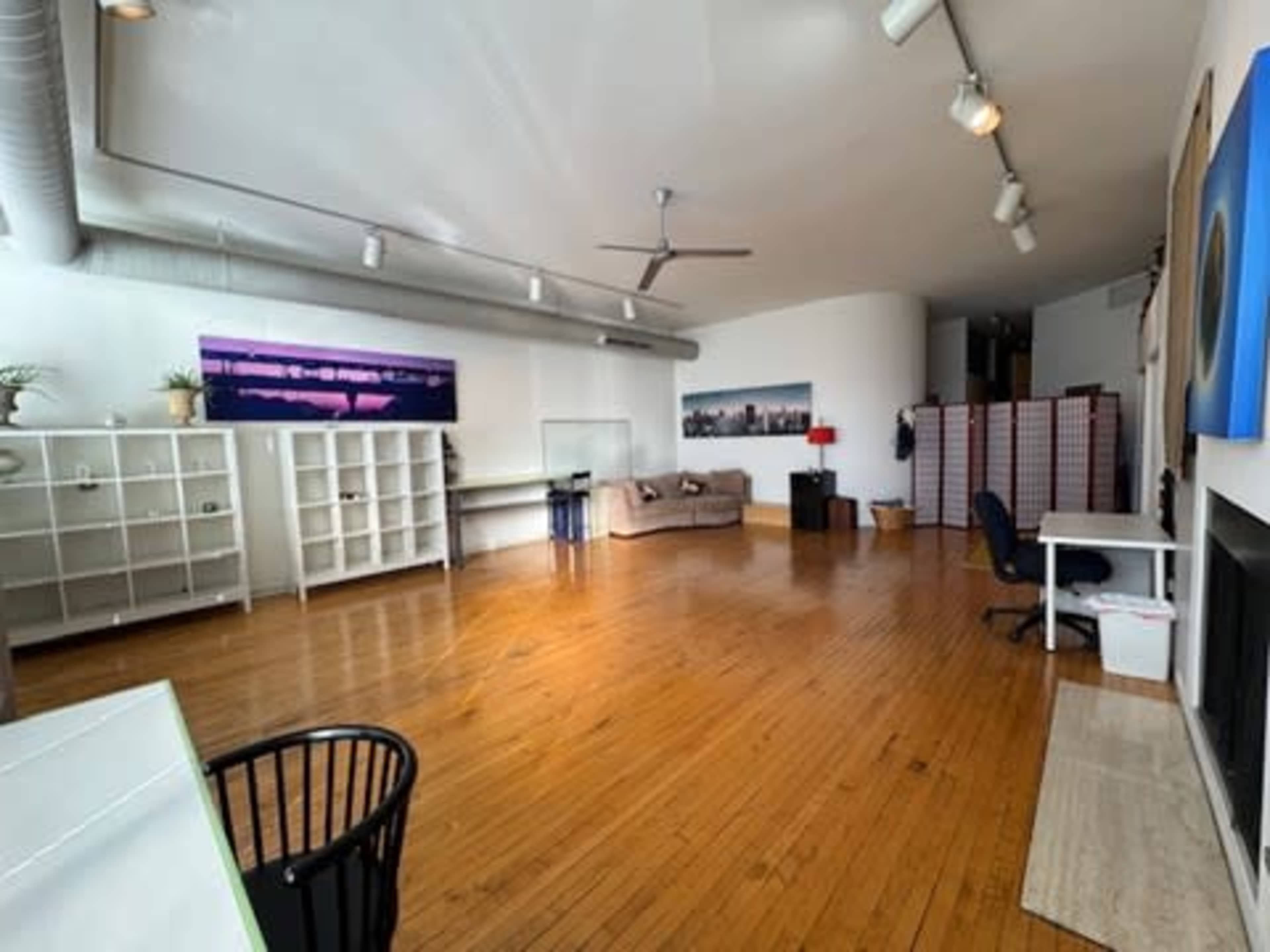 The image shows a spacious, open room featuring wooden floors, shelving units, a couch, a desk, and decorative wall art.