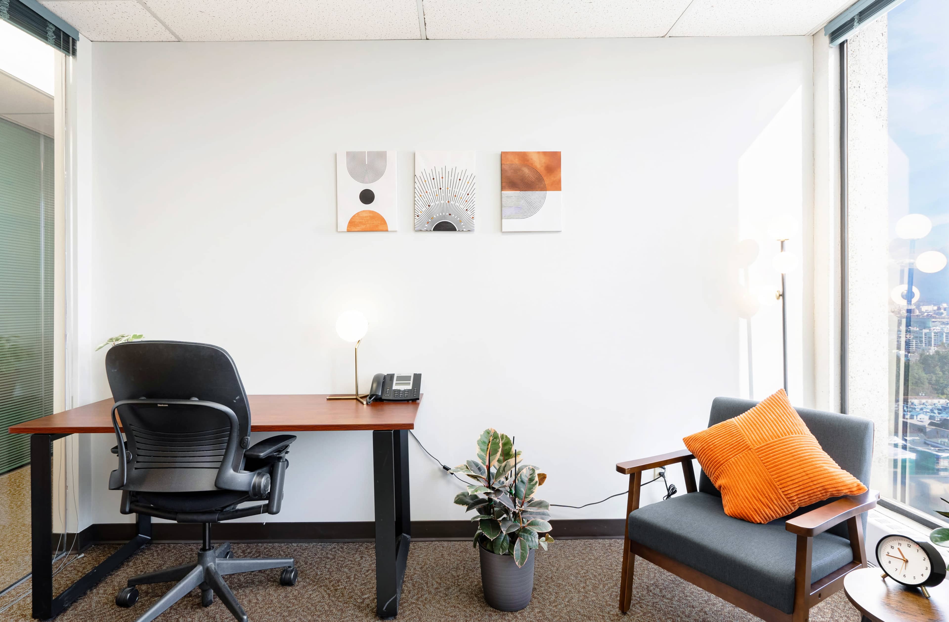 A modern office space featuring a desk with a phone, a chair, artwork on the wall, and a potted plant in a well-lit corner.