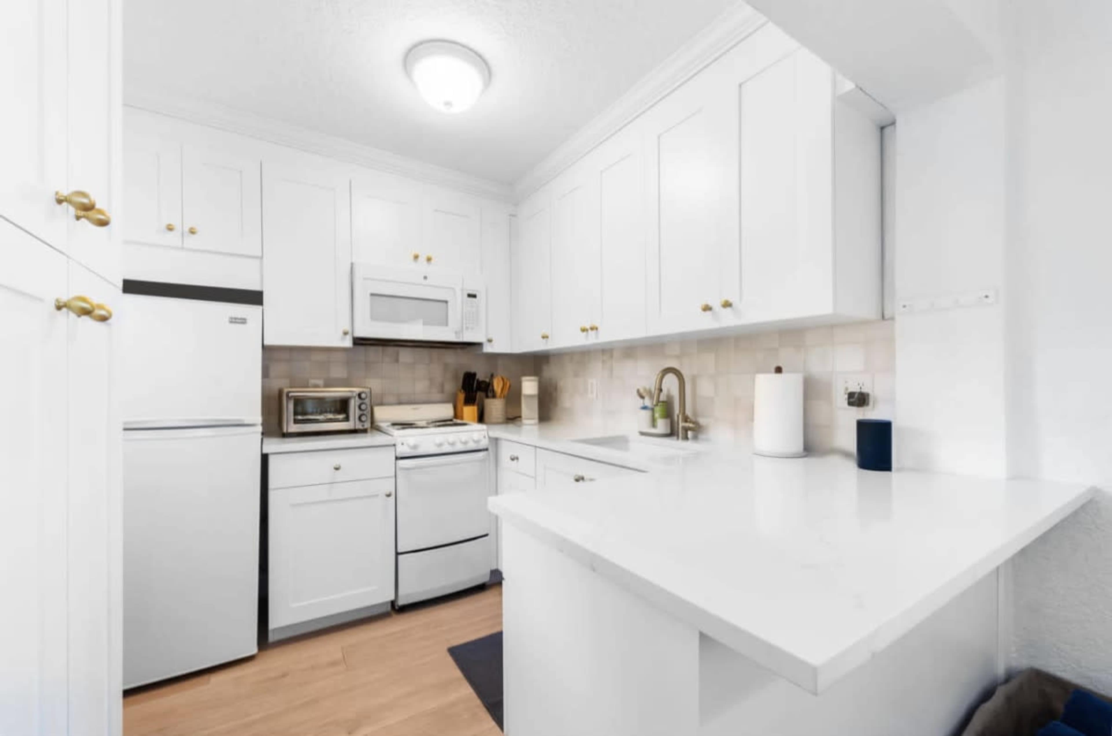 The image shows a compact kitchen with white cabinetry, a stove, a microwave, and a dishwasher, featuring a light countertop and wooden flooring.