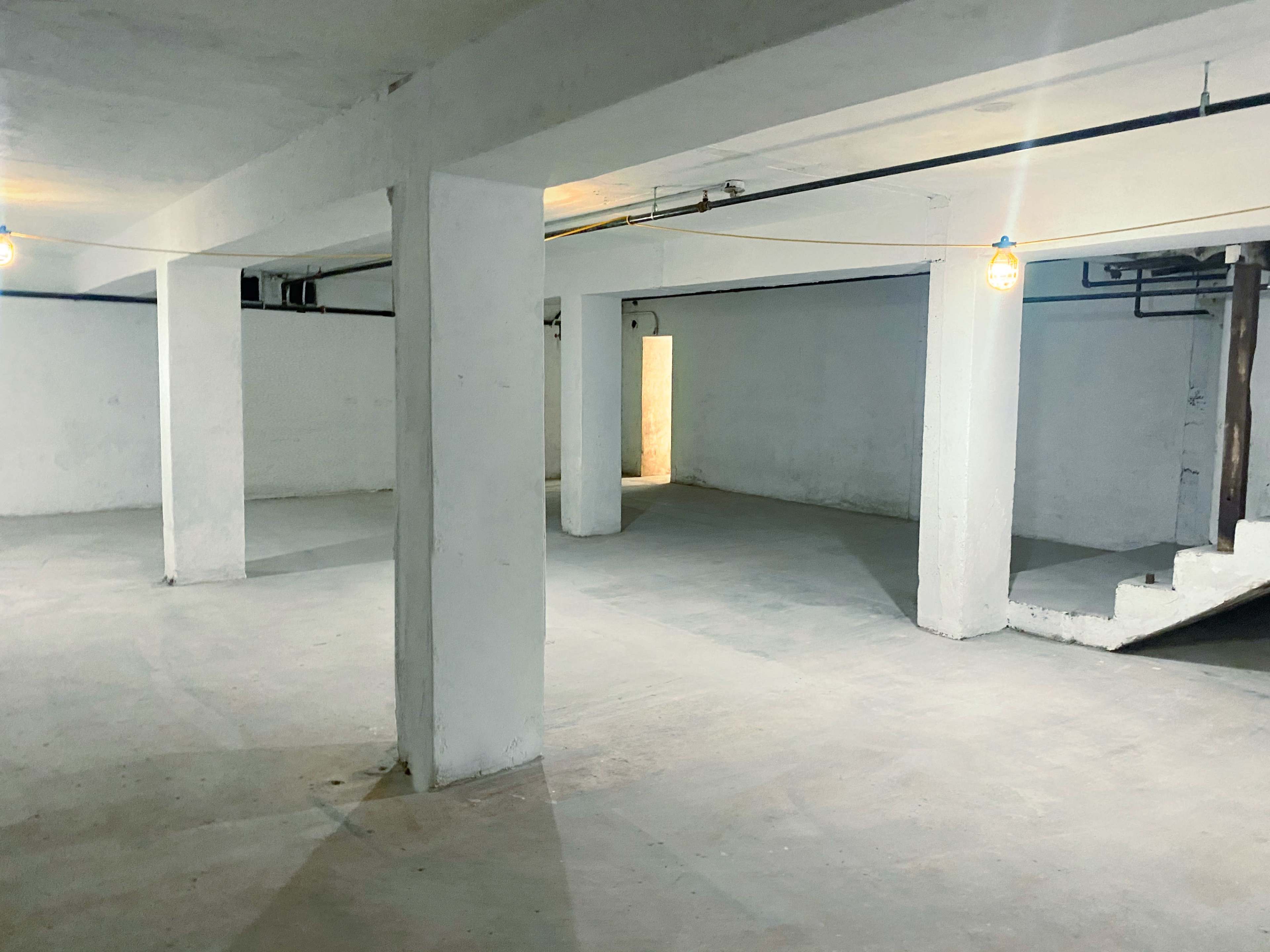A large, empty basement with white walls and concrete floors, illuminated by overhead lights.