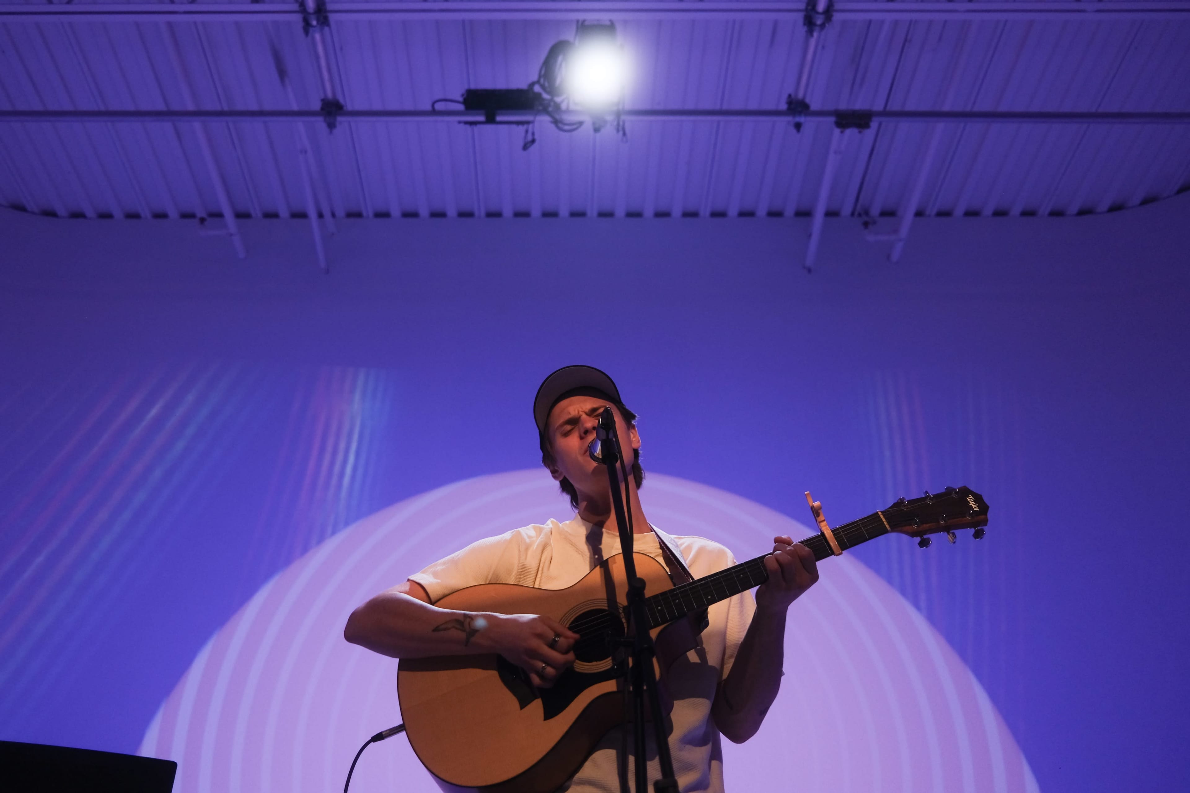 A musician plays an acoustic guitar while singing on stage, with colorful lighting in the background.