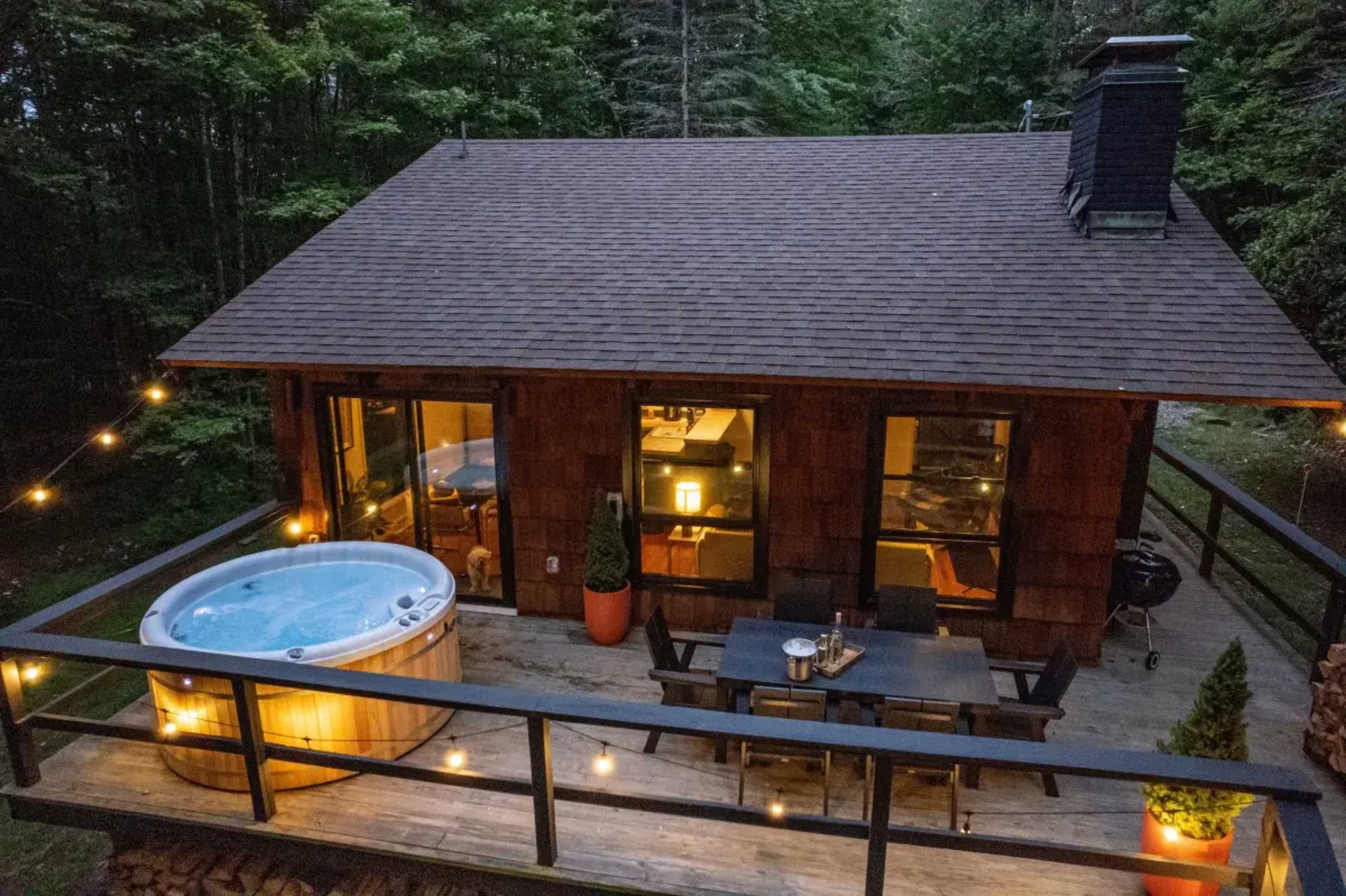 The image shows a wooden cabin with large windows, a hot tub on the deck, and outdoor seating illuminated by string lights, surrounded by trees.
