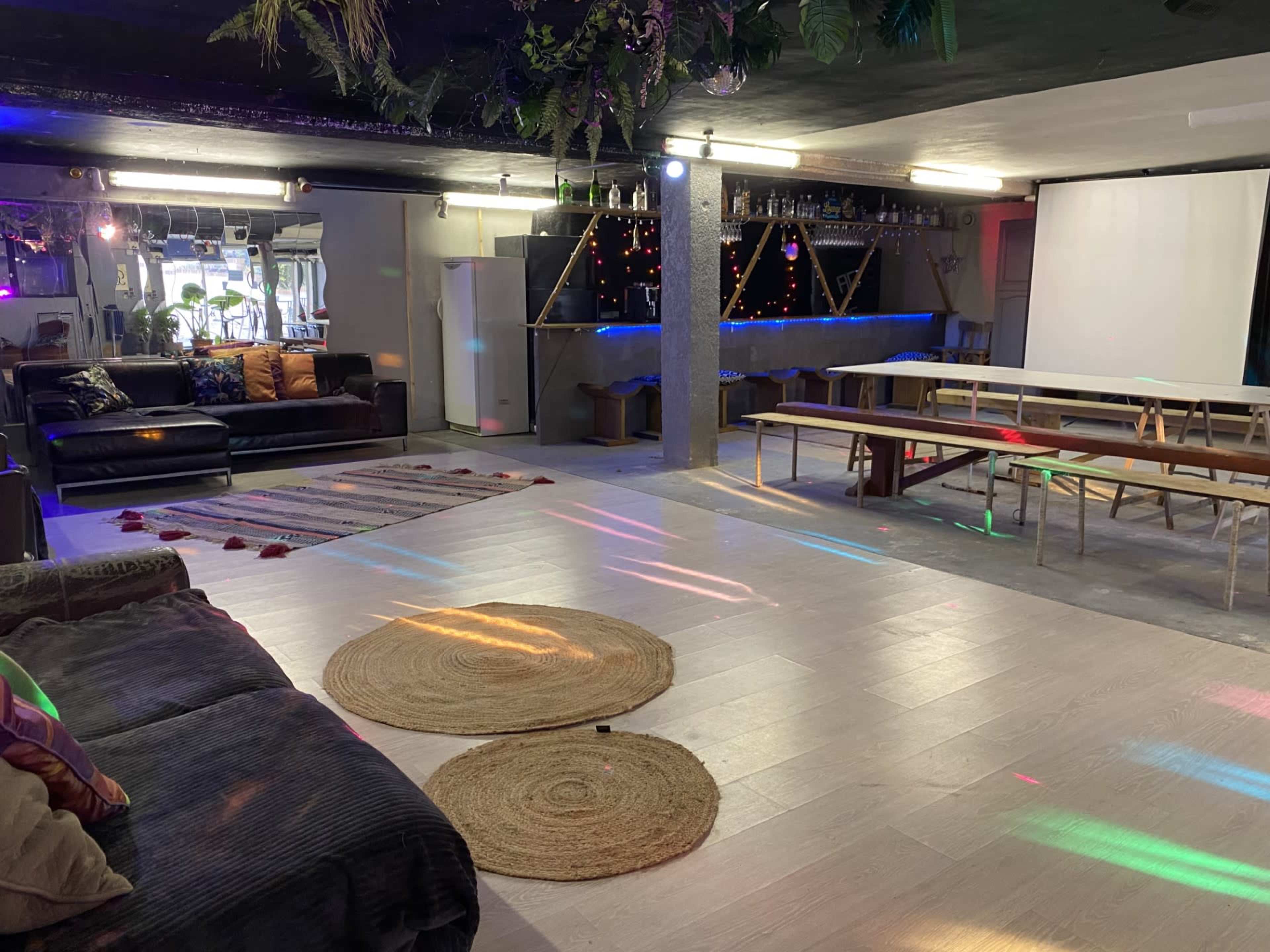 A spacious indoor area with a lounge section featuring sofas, a bar with shelves of bottles, and a table setup, illuminated by colorful lights.
