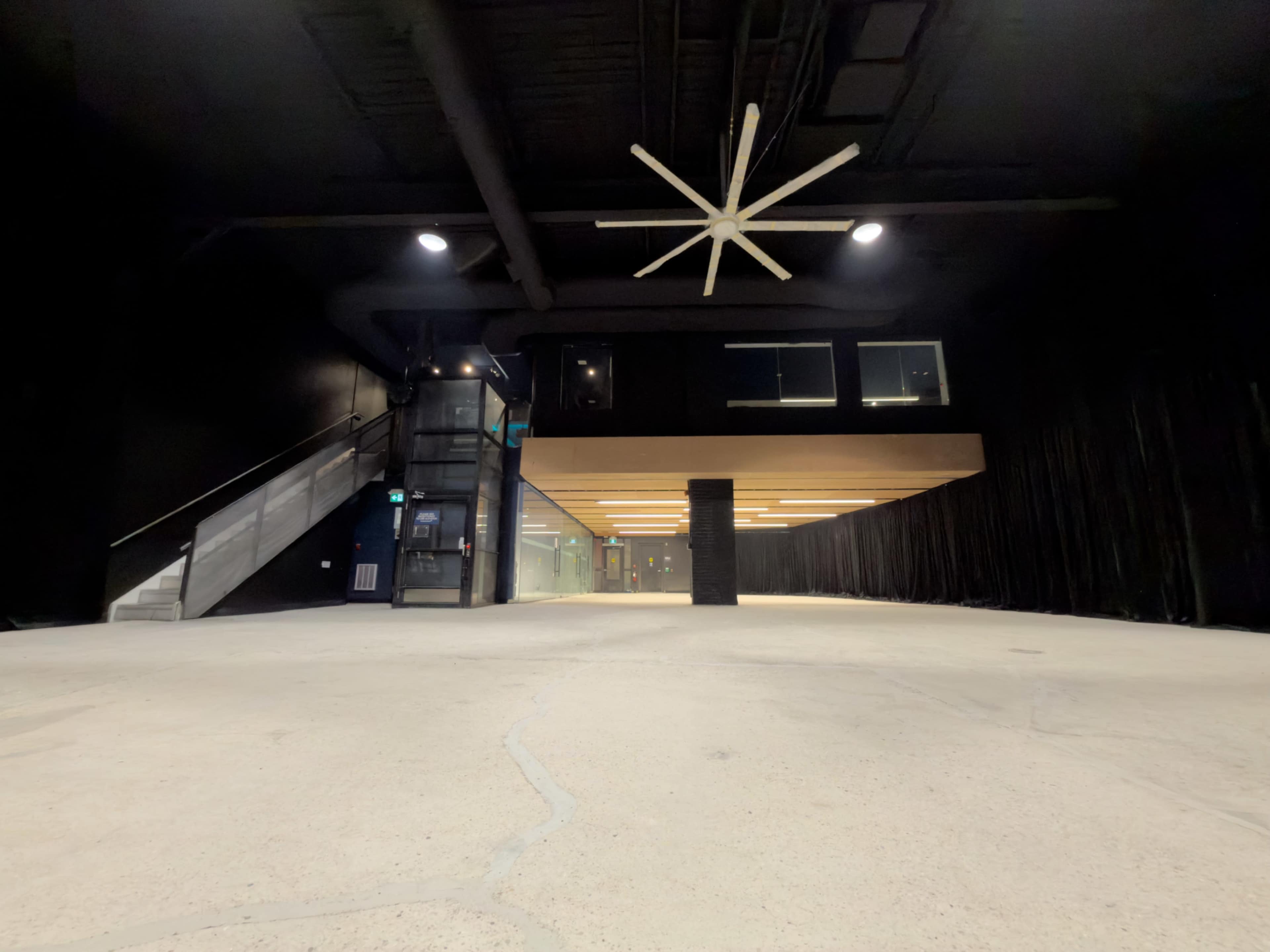 The image shows a large, mostly empty room with black walls and a ceiling fan, featuring a staircase on one side and a balcony above.