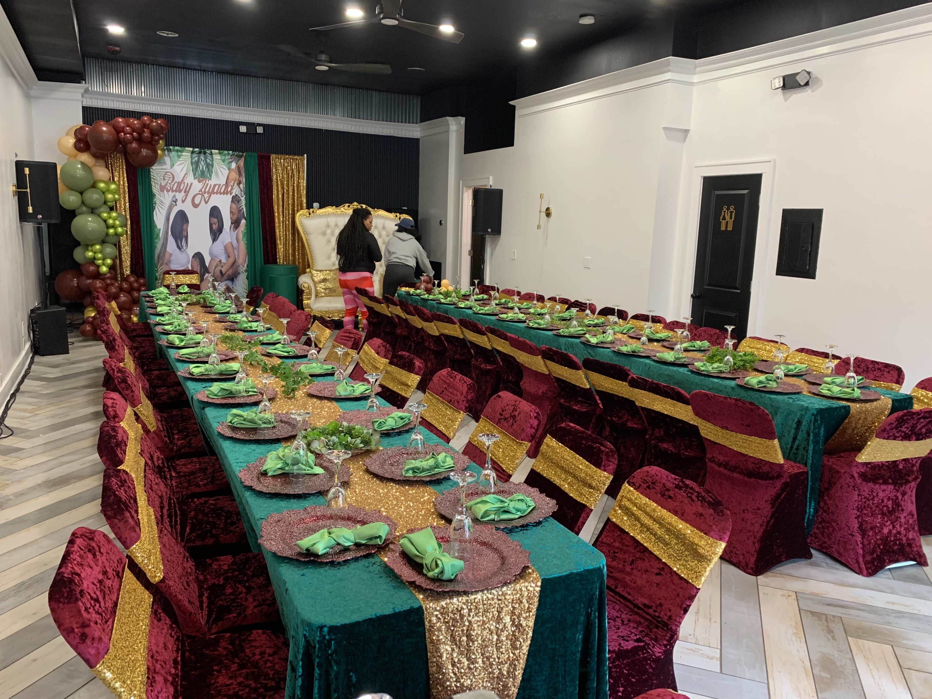 A large event space is set up with two long tables covered in green and maroon tablecloths, adorned with gold accents and neatly arranged place settings.