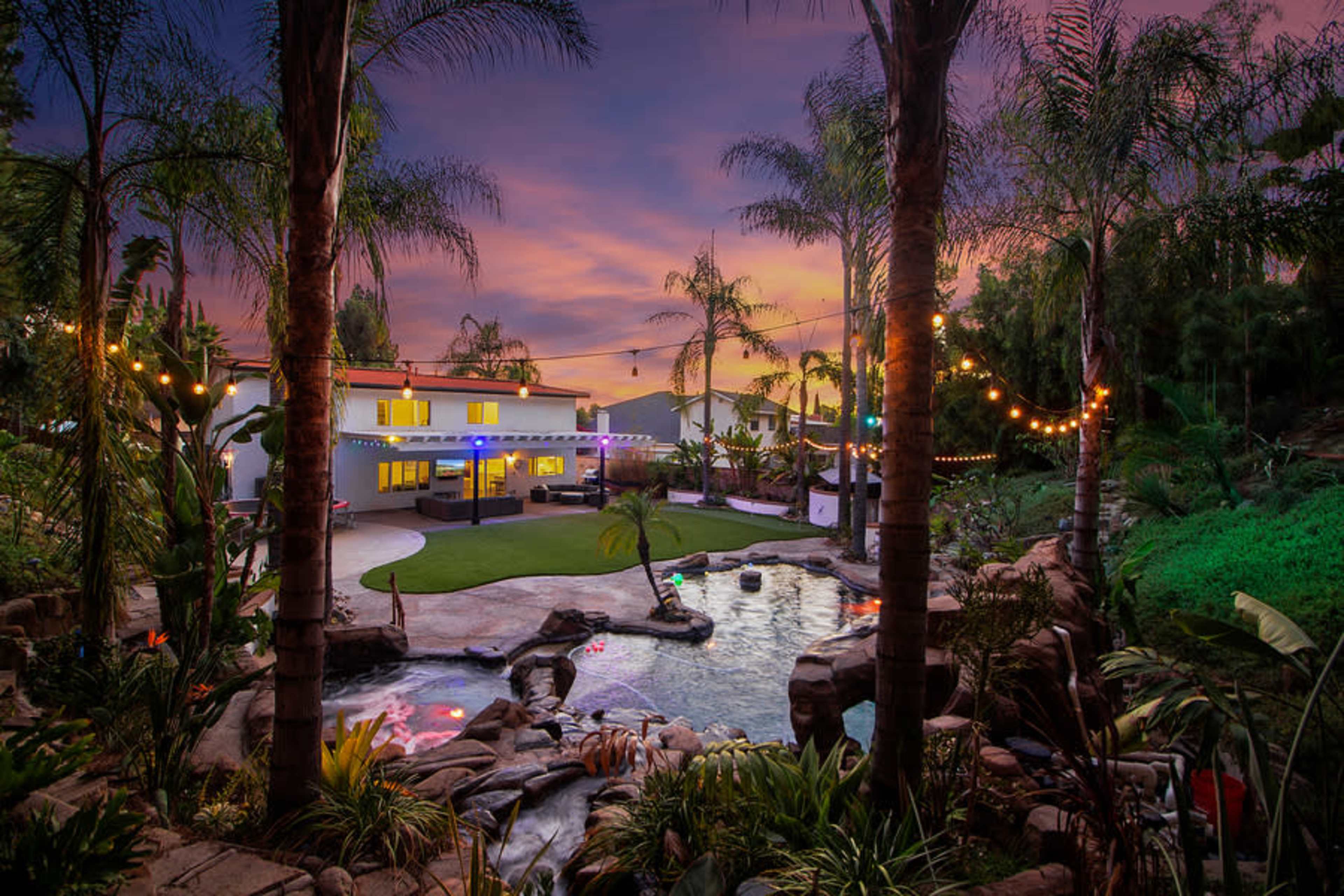 A landscaped backyard features a swimming pool, palm trees, and string lights, set against a colorful sunset sky.