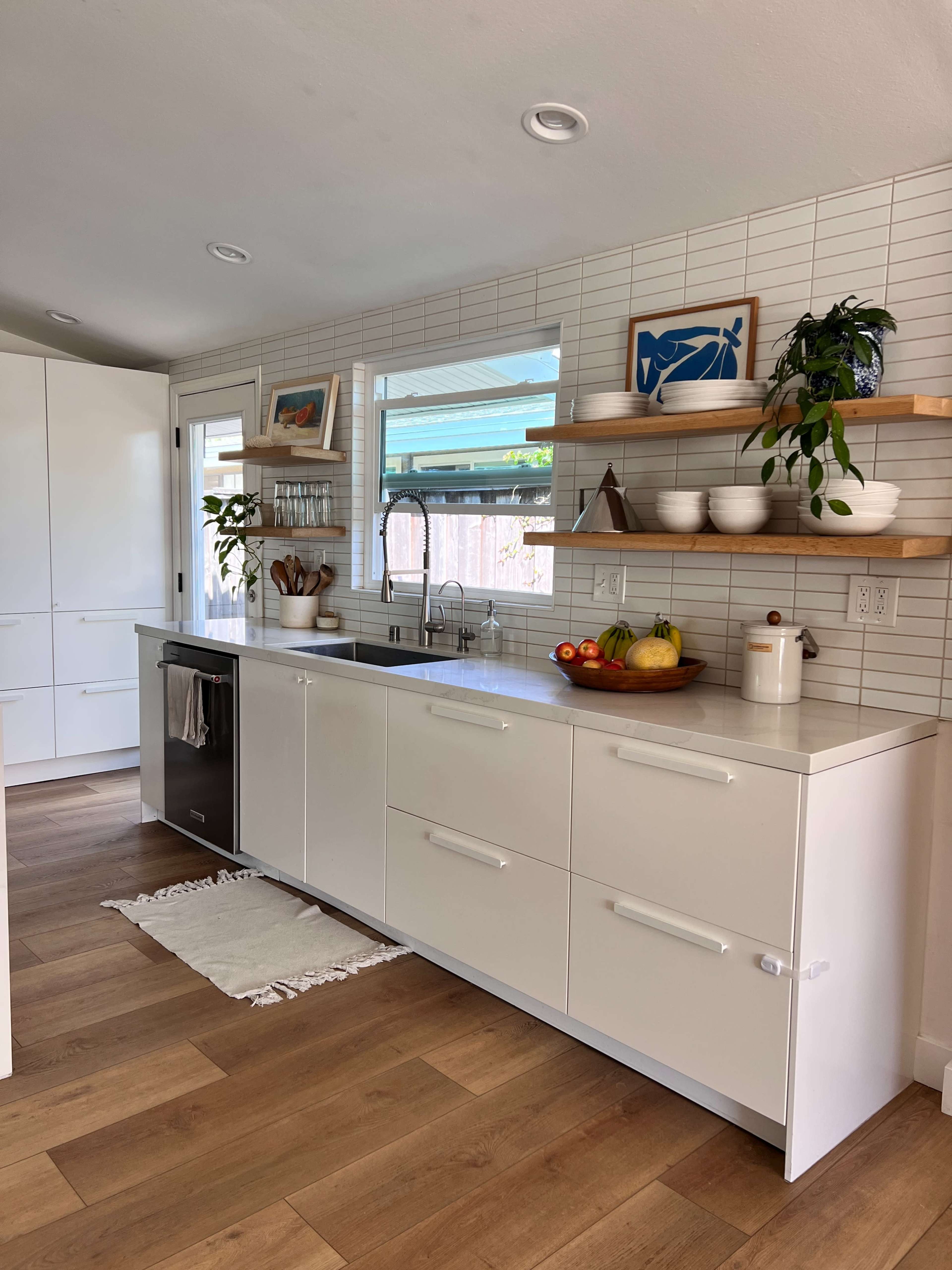The image shows a modern kitchen with white cabinetry, wooden shelves holding decorative items and plants, and a countertop with fresh fruit and a sink.