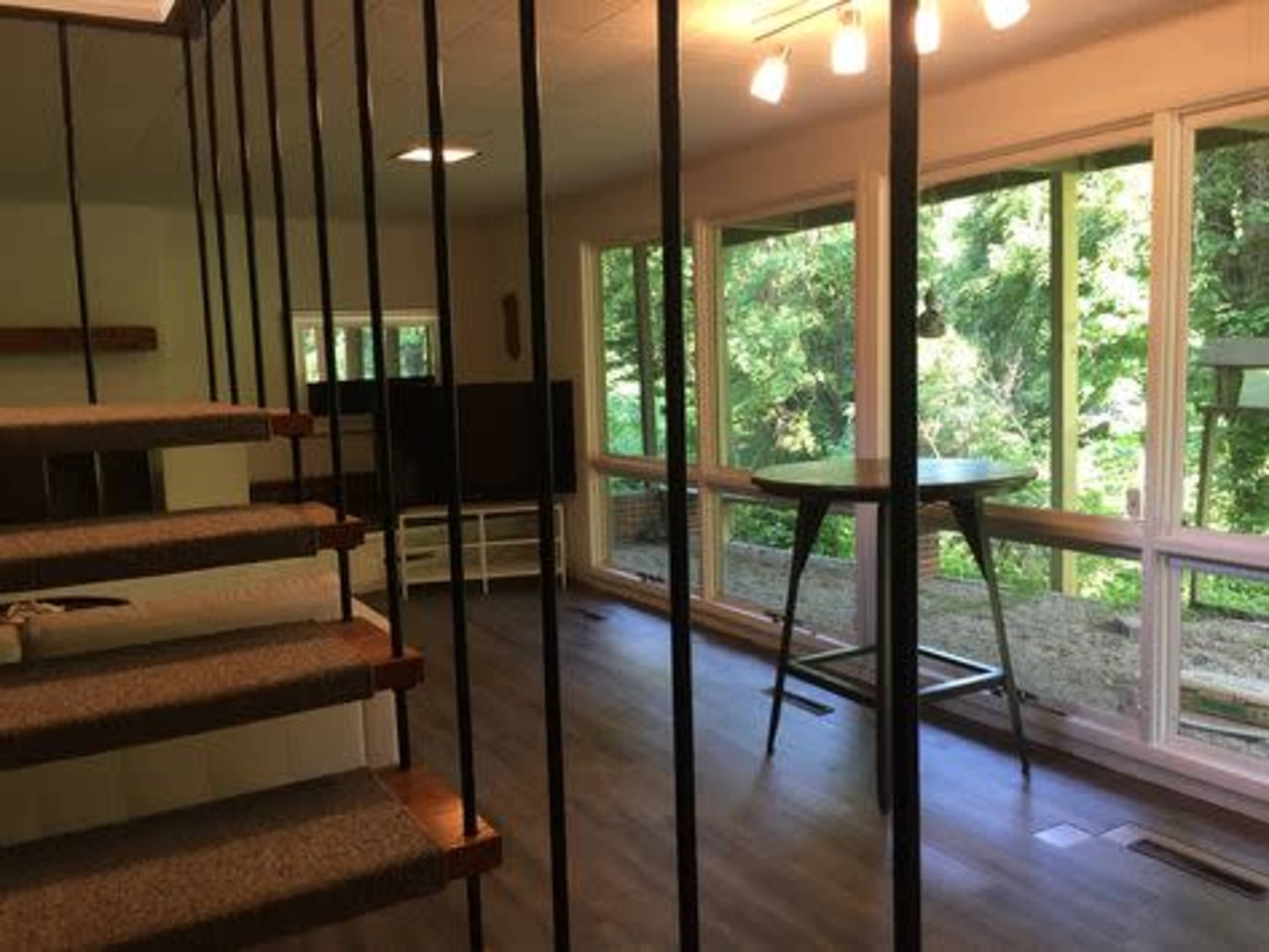 The image shows a modern interior space with a staircase, large windows overlooking greenery, and a small table set against the glass.