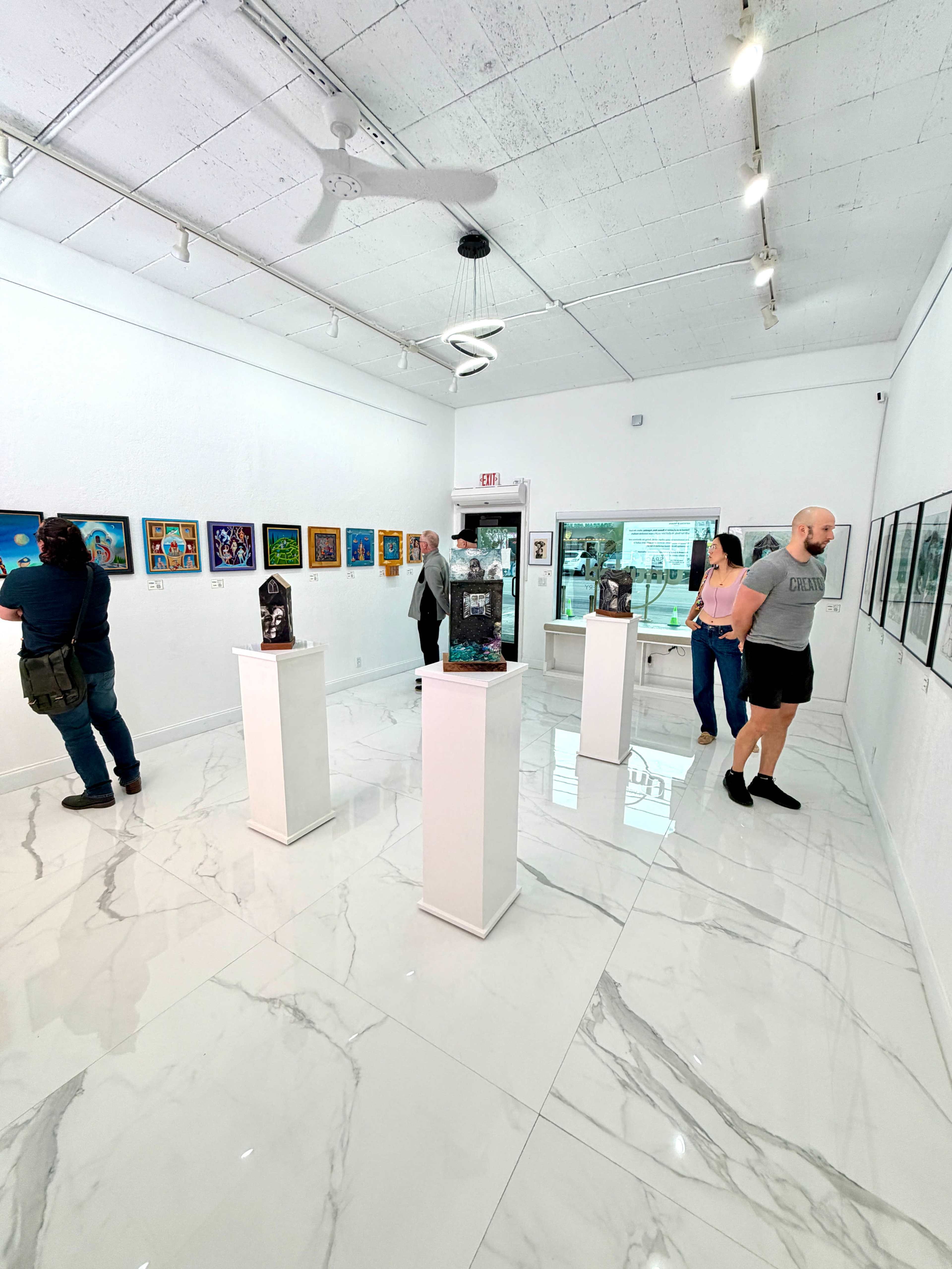 The image shows an art gallery with white walls, marble flooring, and several artworks displayed on the walls and on pedestals.