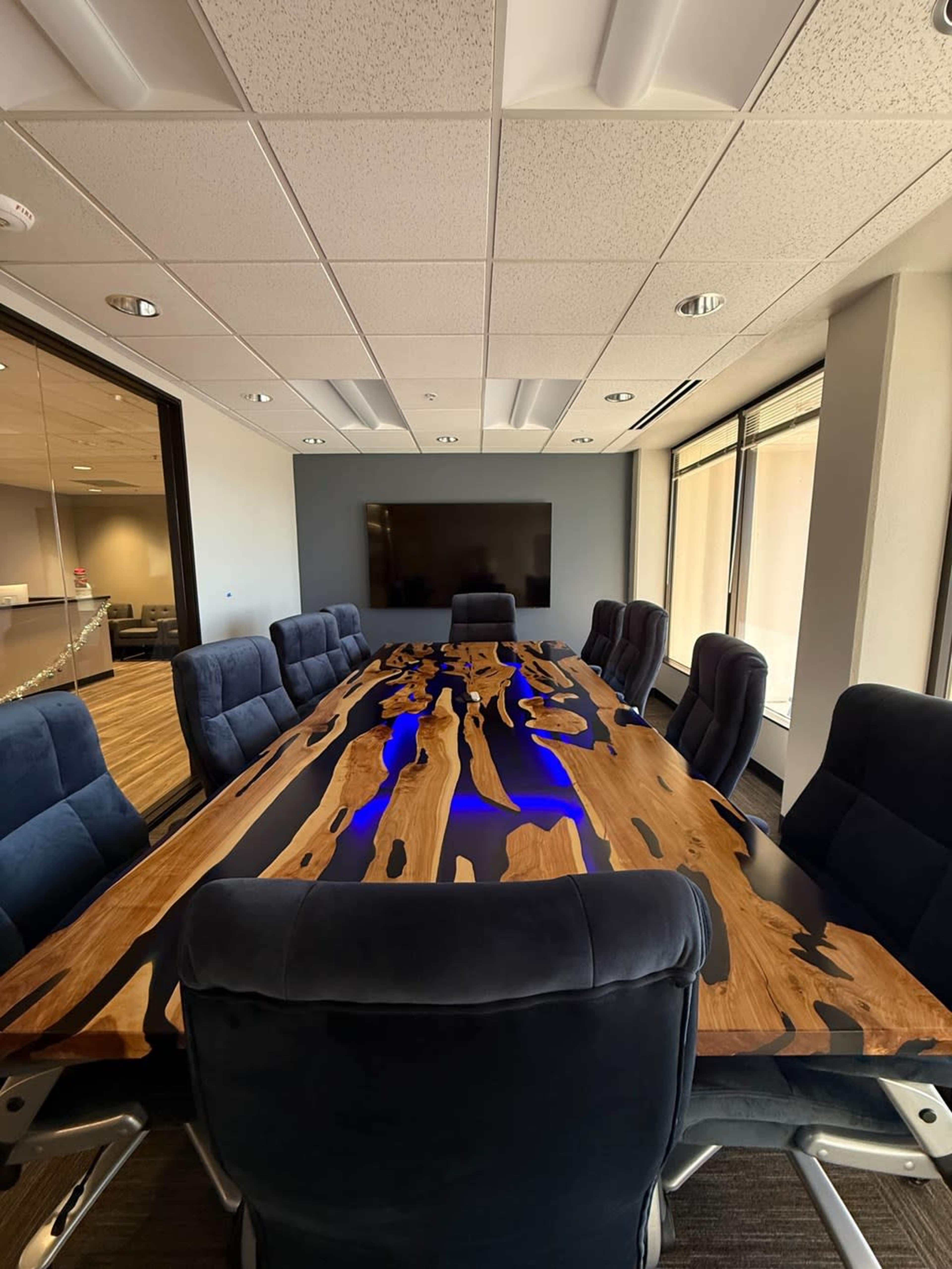 A modern conference room features a large wooden table with a sleek design, surrounded by high-backed chairs and a wall-mounted screen.