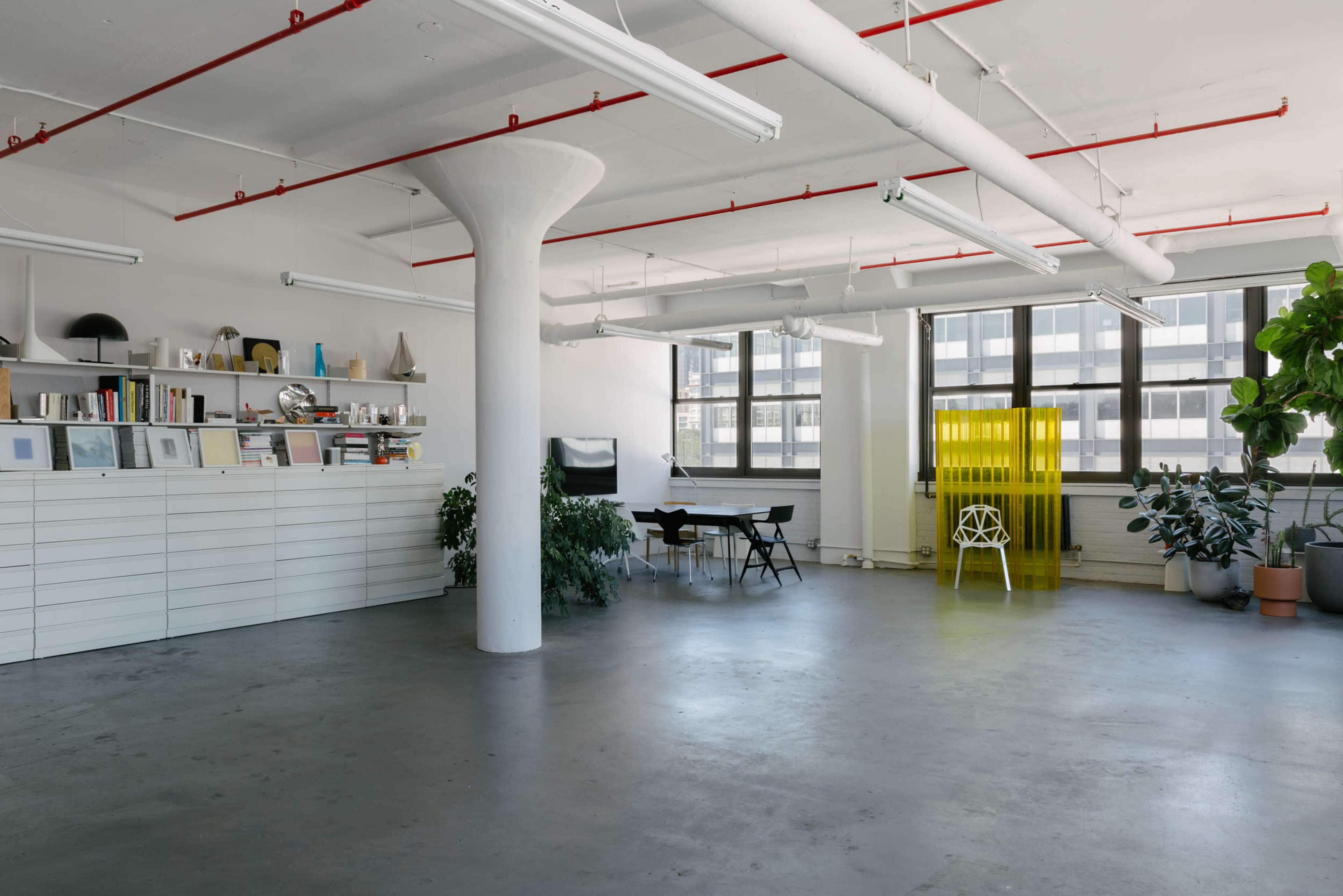 The image shows a spacious, minimalist office interior with large windows, a central pillar, and various plants, furniture, and shelving units.