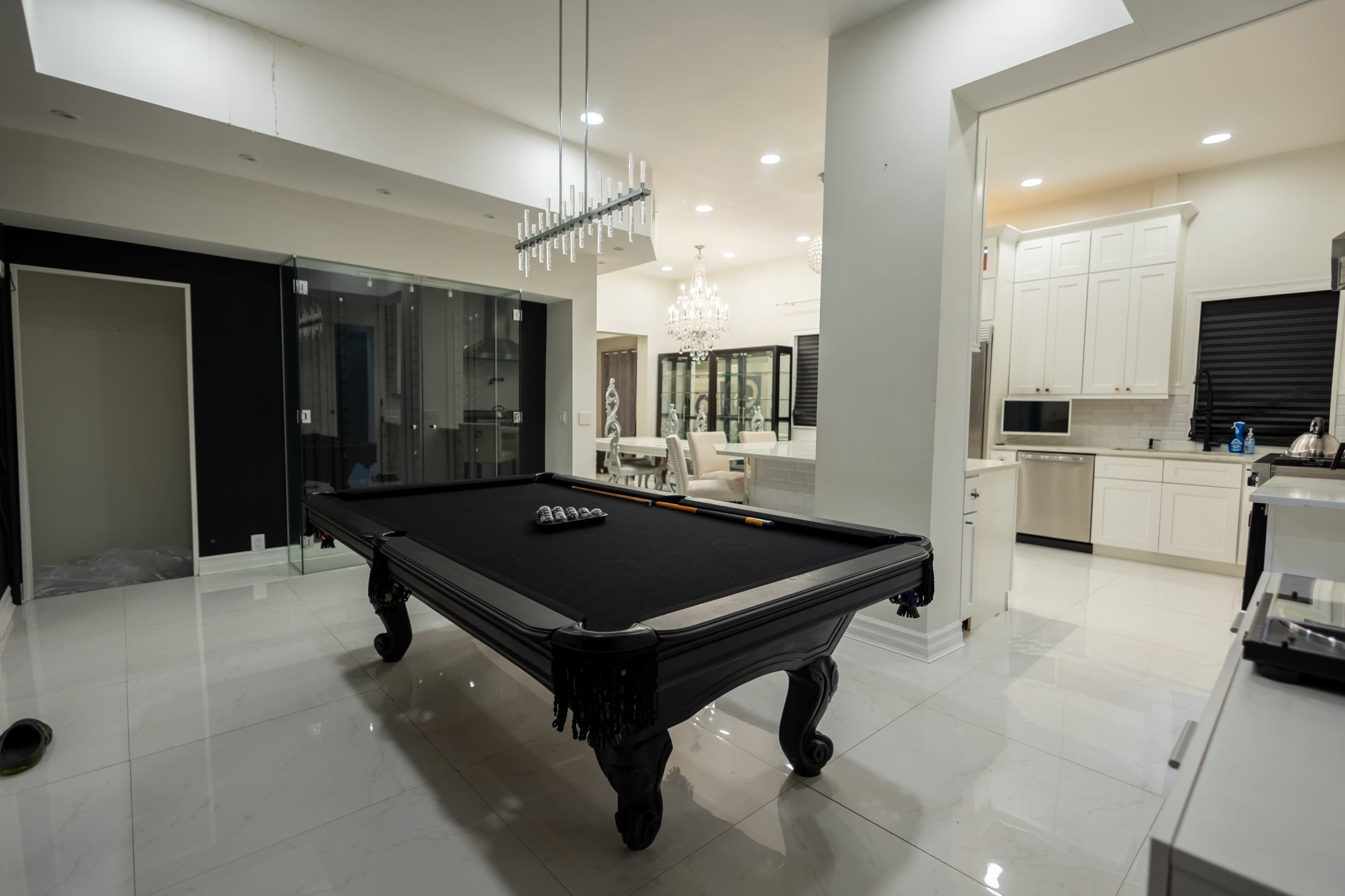 The image shows a modern interior featuring a black billiard table in a well-lit room with white tile flooring and an open kitchen area.
